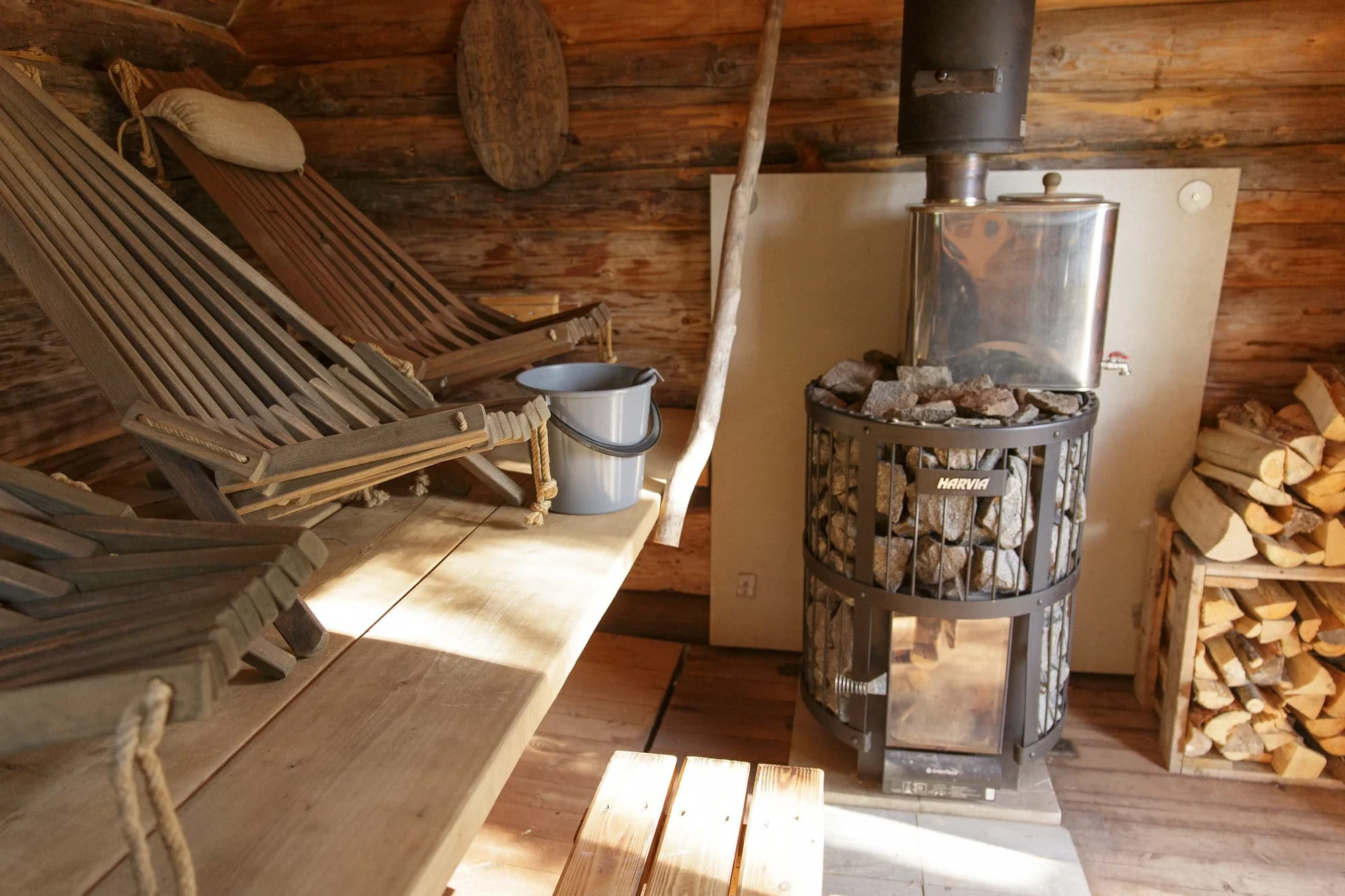 Wooden interior with a wood-burning stove, firewood, and wooden furniture, including chairs and a table.