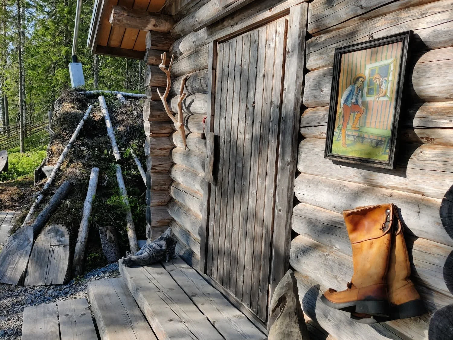 The exterior corner of a rustic wooden cabin with a closed wooden door, a painting on the wall depicting two people in a room, a mounted deer antler, a pair of tan leather boots, and a blurry view of trees and a fence in the background.