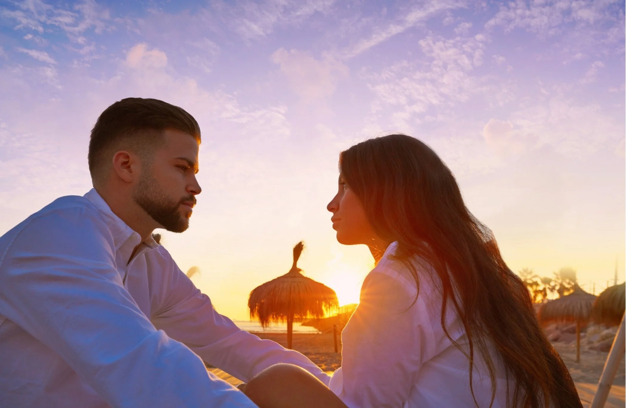 A man and woman sitting face to face on a beach at sunset, looking at each other, with thatched umbrellas and a pink and purple sky in the background.