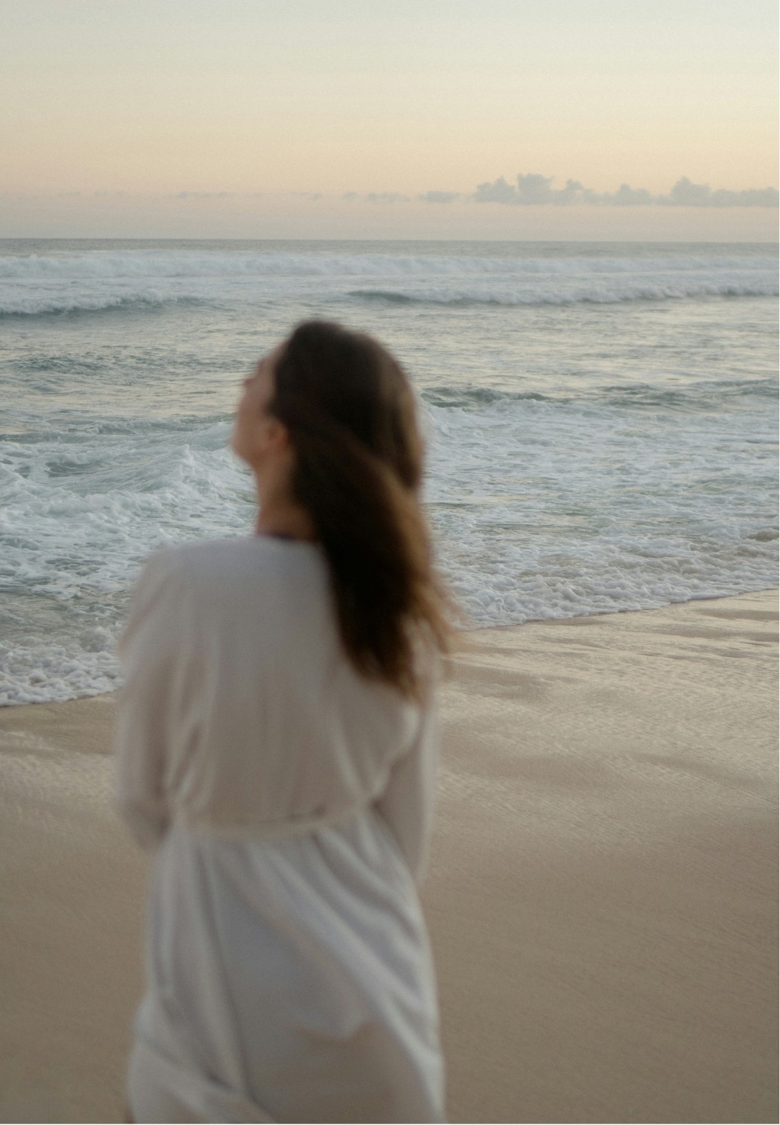 A woman in a white dress standing on a sandy beach, looking out at the ocean during sunset.
