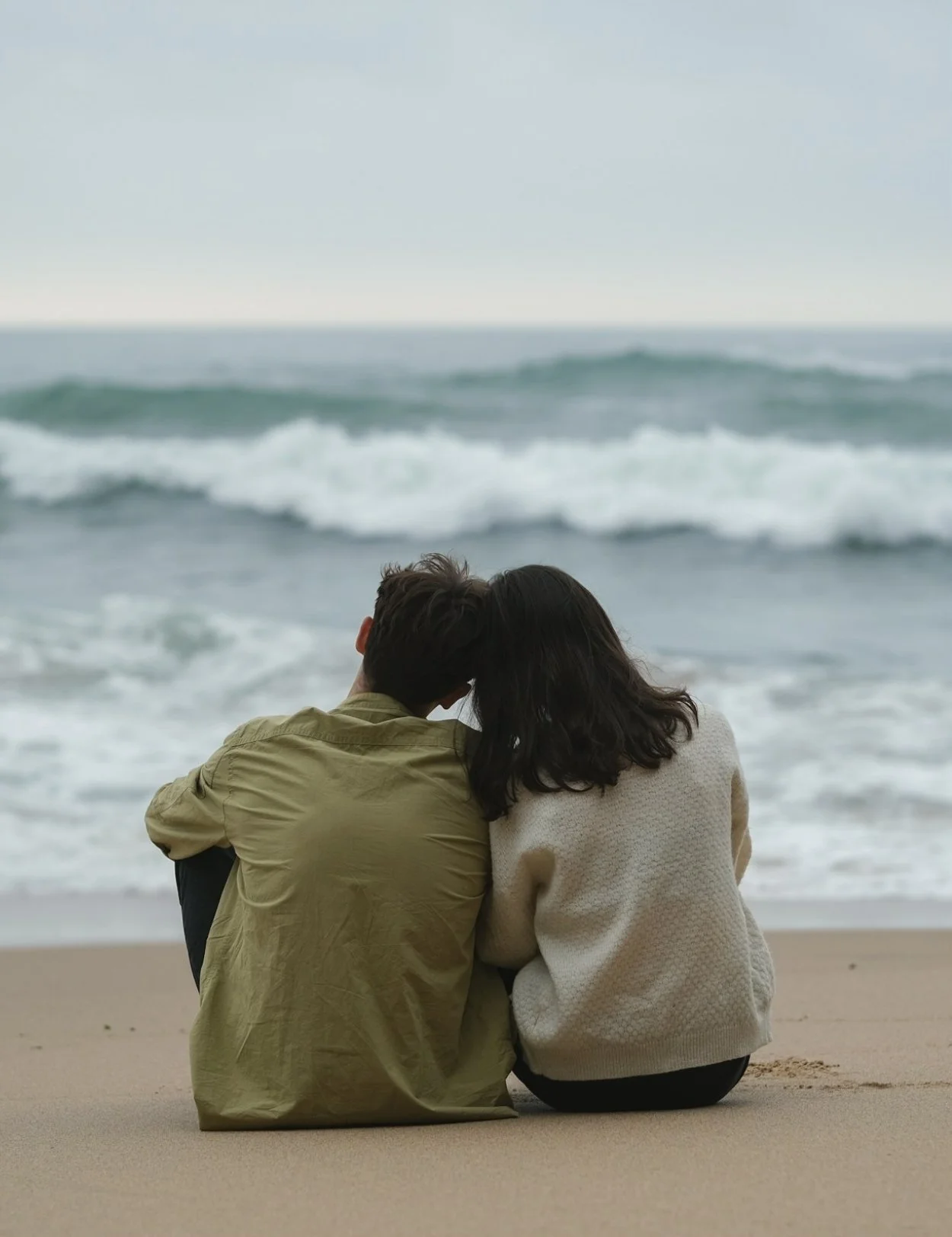 A couple sitting on the beach, facing the ocean, with their heads close together.
