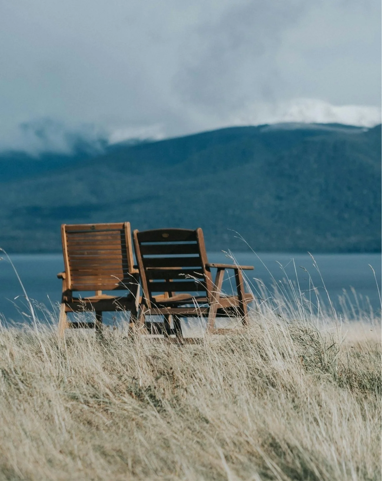 Two wooden chairs on a grassy area near a body of water with mountains and cloudy sky in the background.