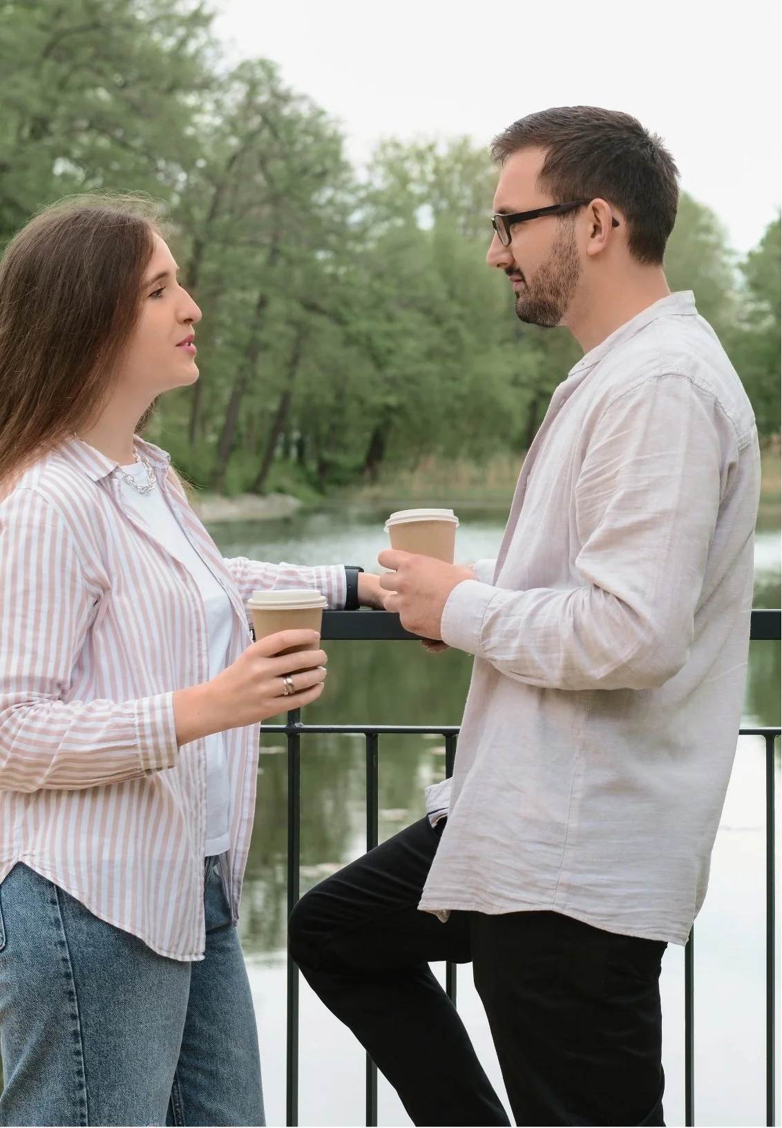 A man and woman standing outdoors near a body of water, holding coffee cups and engaging in conversation, with trees in the background.