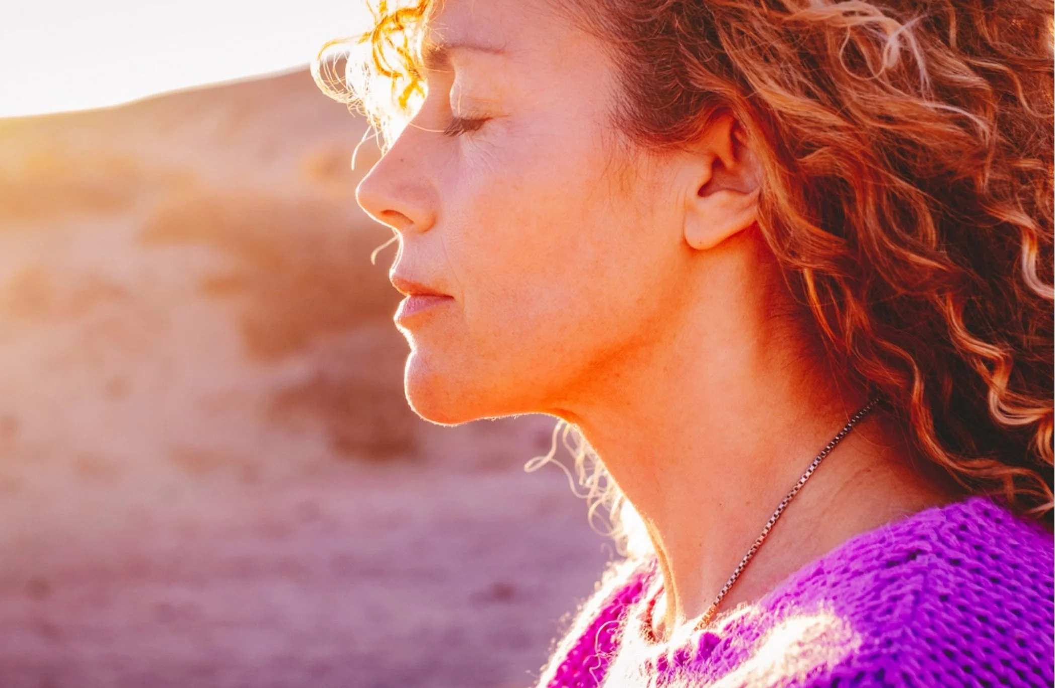 Profile of a woman with curly red hair with eyes closed, glowing sunlight in the background.
