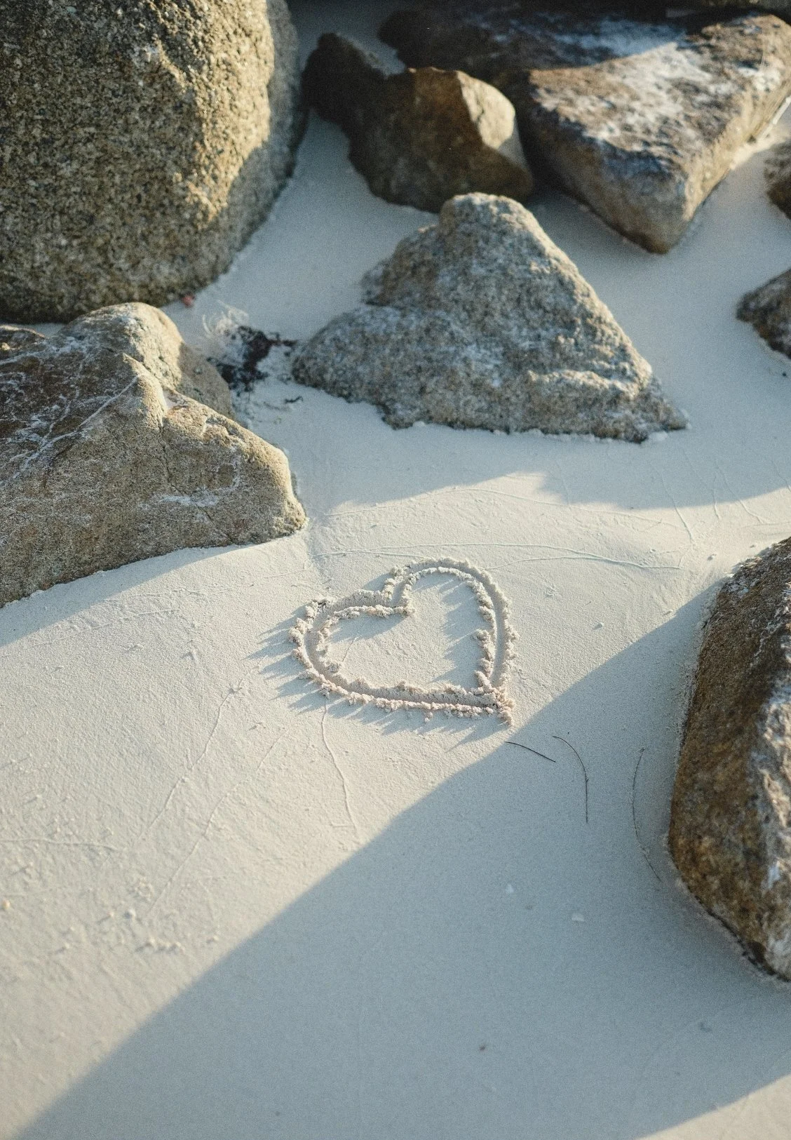 A heart shape drawn in white sand with small rocks around it, on a sandy surface near some large rocks.