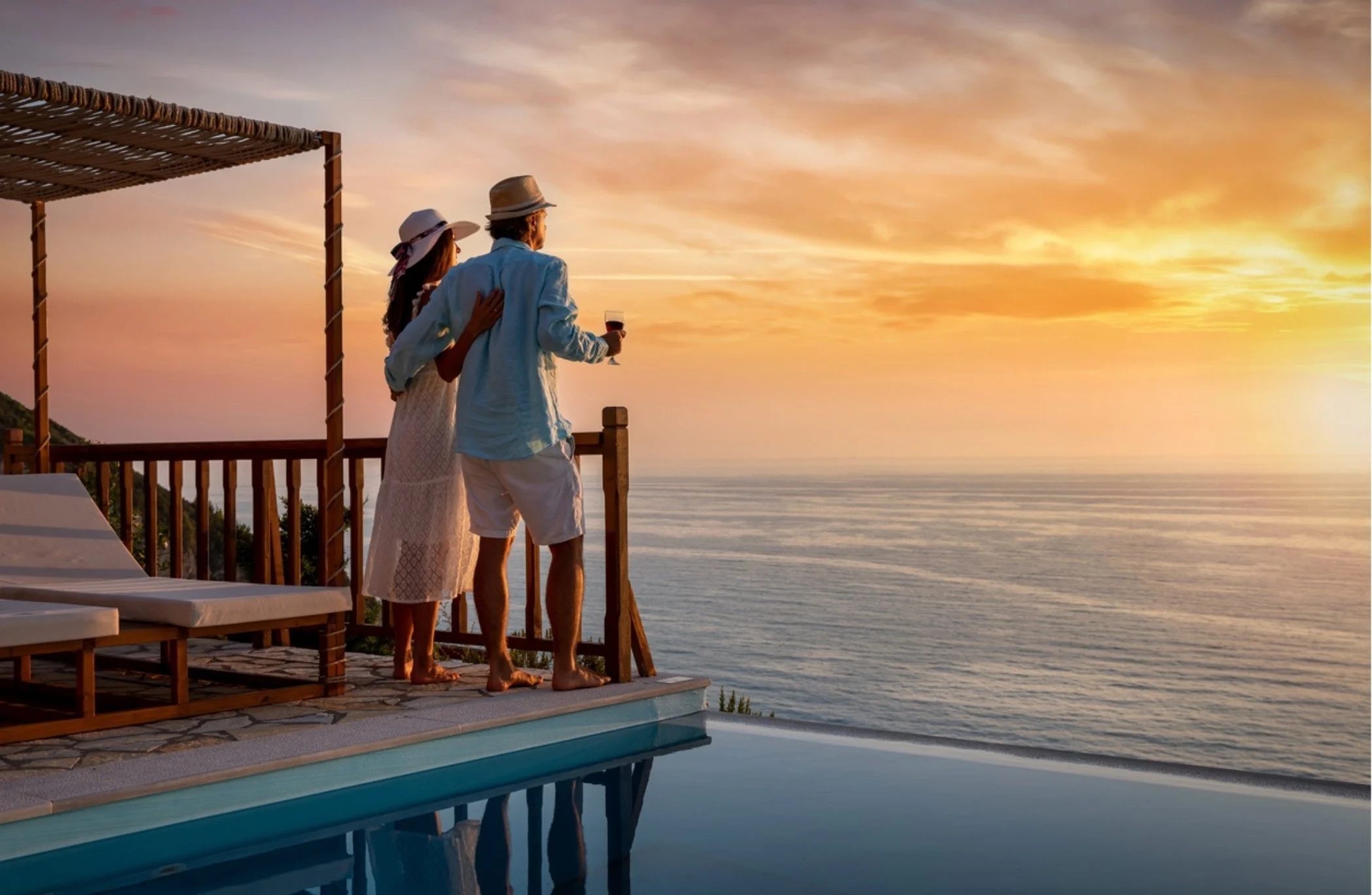 A couple standing on a balcony by a pool, embracing and holding glasses of wine, watching the sunset over the ocean.