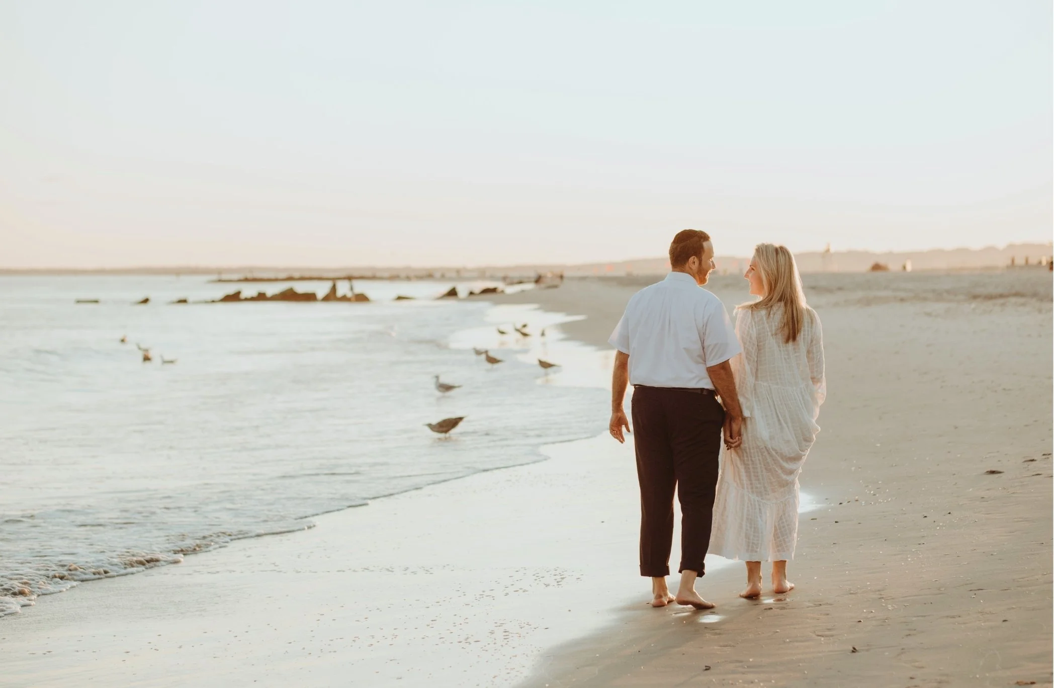 A couple walking on the beach at sunset, holding hands and smiling at each other.
