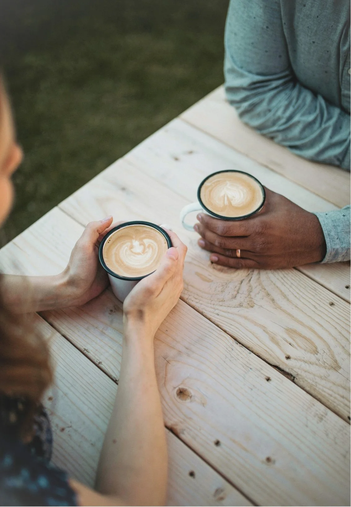 Two people sitting at a wooden table, holding cups of coffee with latte art, outside or in a well-lit area.