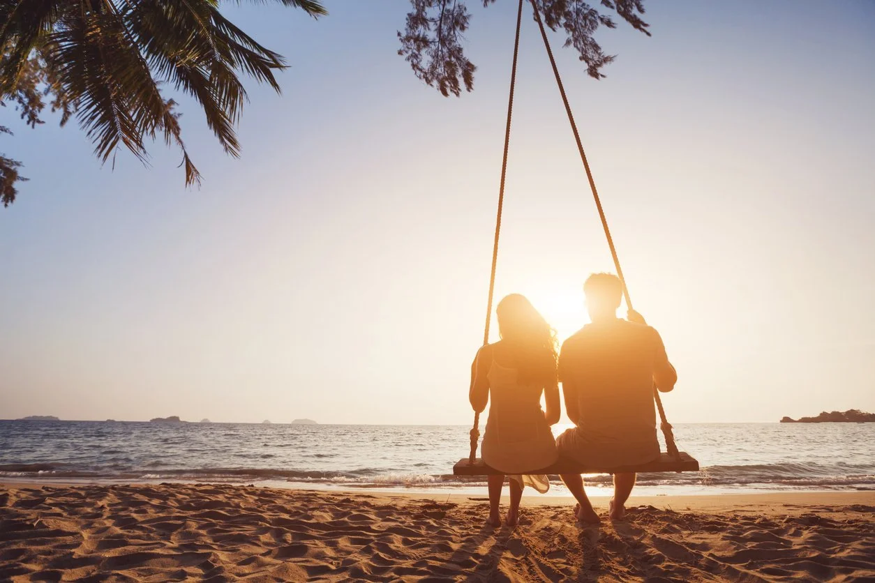 A couple sitting on a swing on the beach during sunset, with palm tree branches above and the ocean in the background.