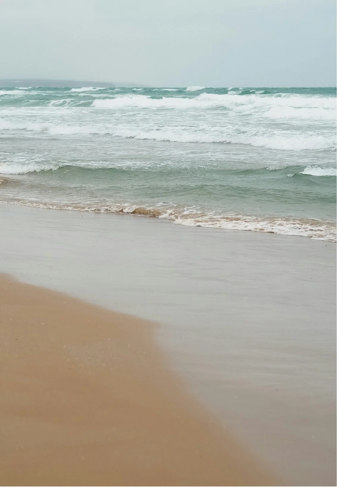 Overcast sky over a sandy beach with gentle waves hitting the shore.