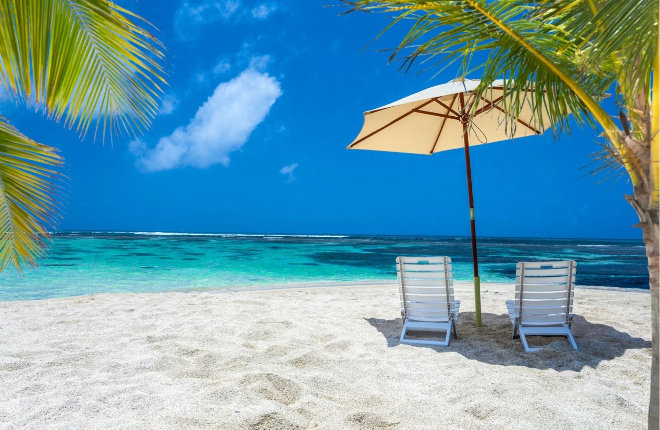 A tropical beach scene with white sand, two white lounge chairs, a beige umbrella, and palm trees at the shoreline. The ocean is turquoise with gentle waves, and the sky is blue with some clouds.