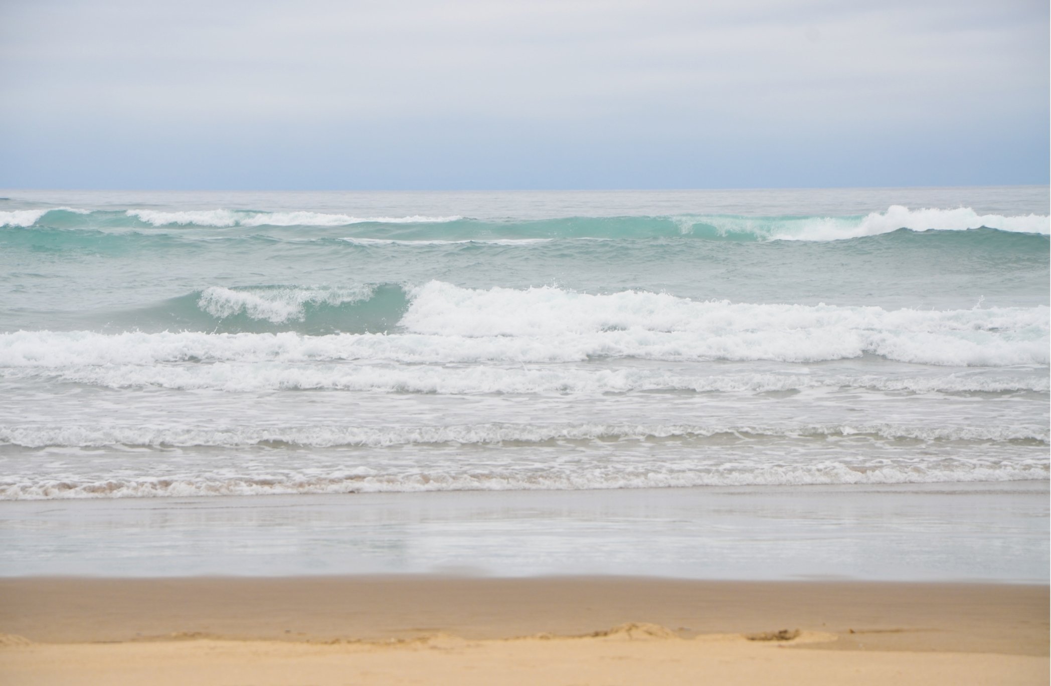 Beach scene with sand in the foreground, and waves and ocean in the background under a cloudy sky.