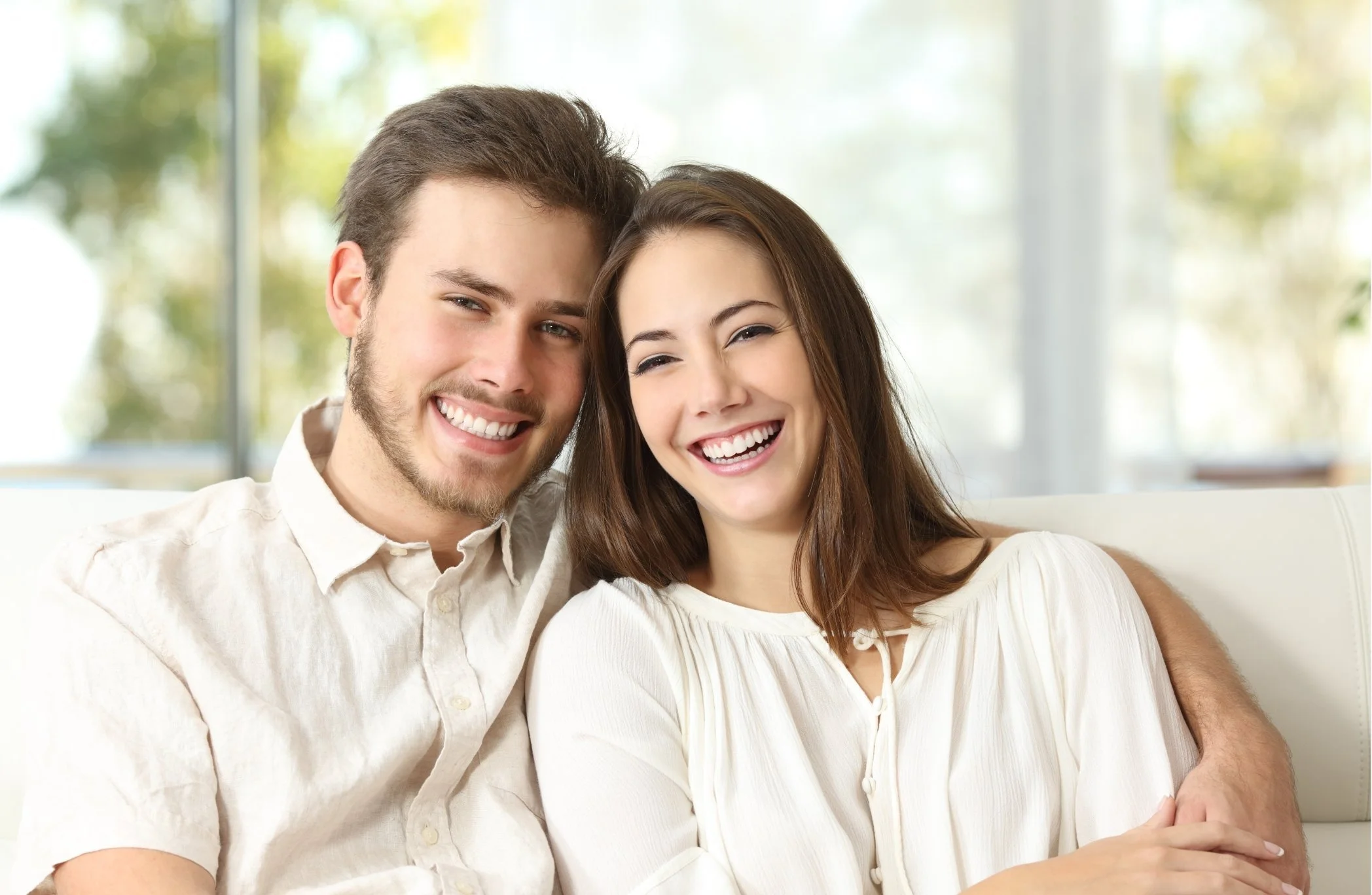 A smiling young man and woman with their heads close together, sitting on a sofa in a bright room.