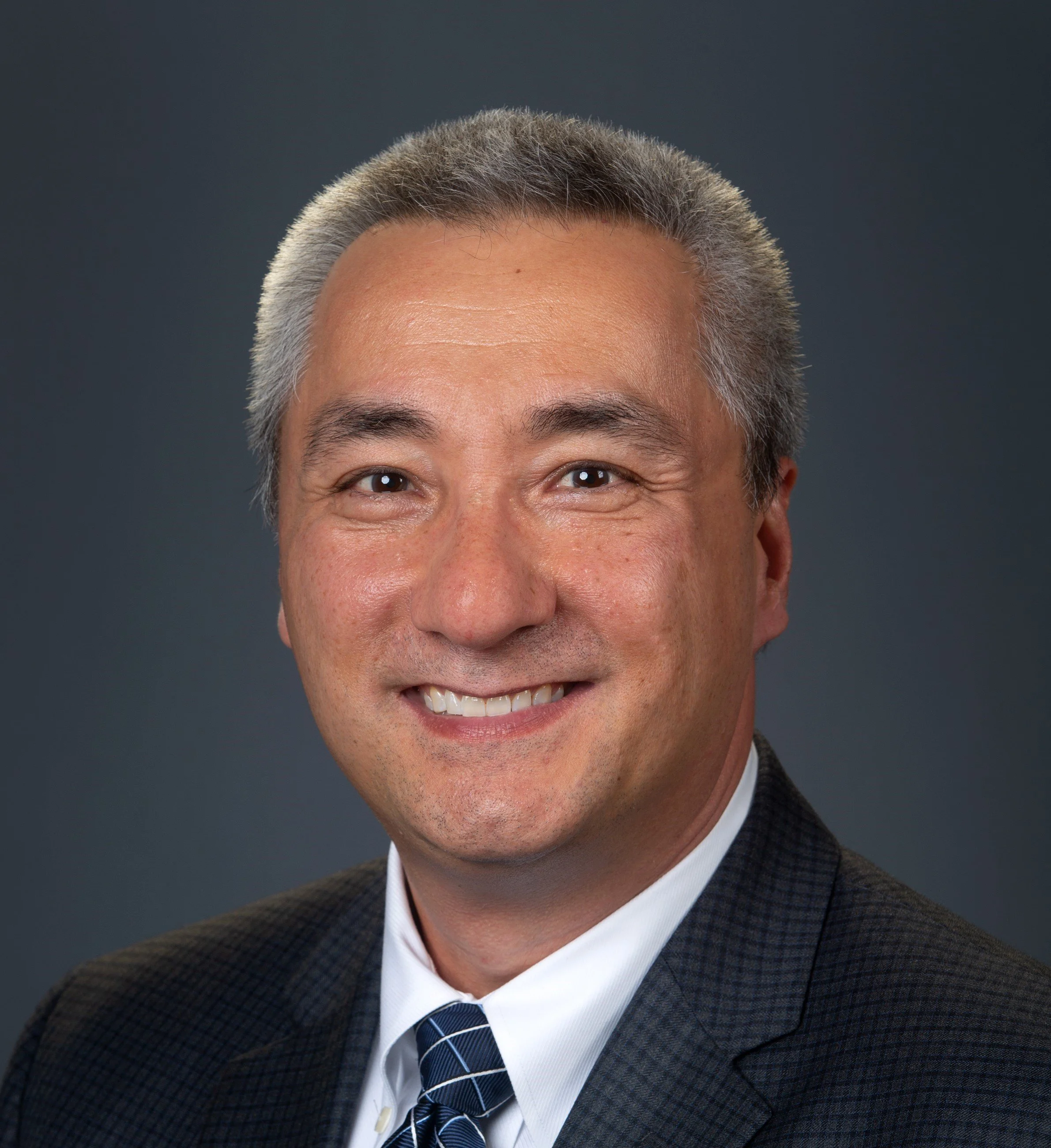 Headshot of a middle-aged man with short gray hair, smiling, wearing a suit and tie against a plain dark background.