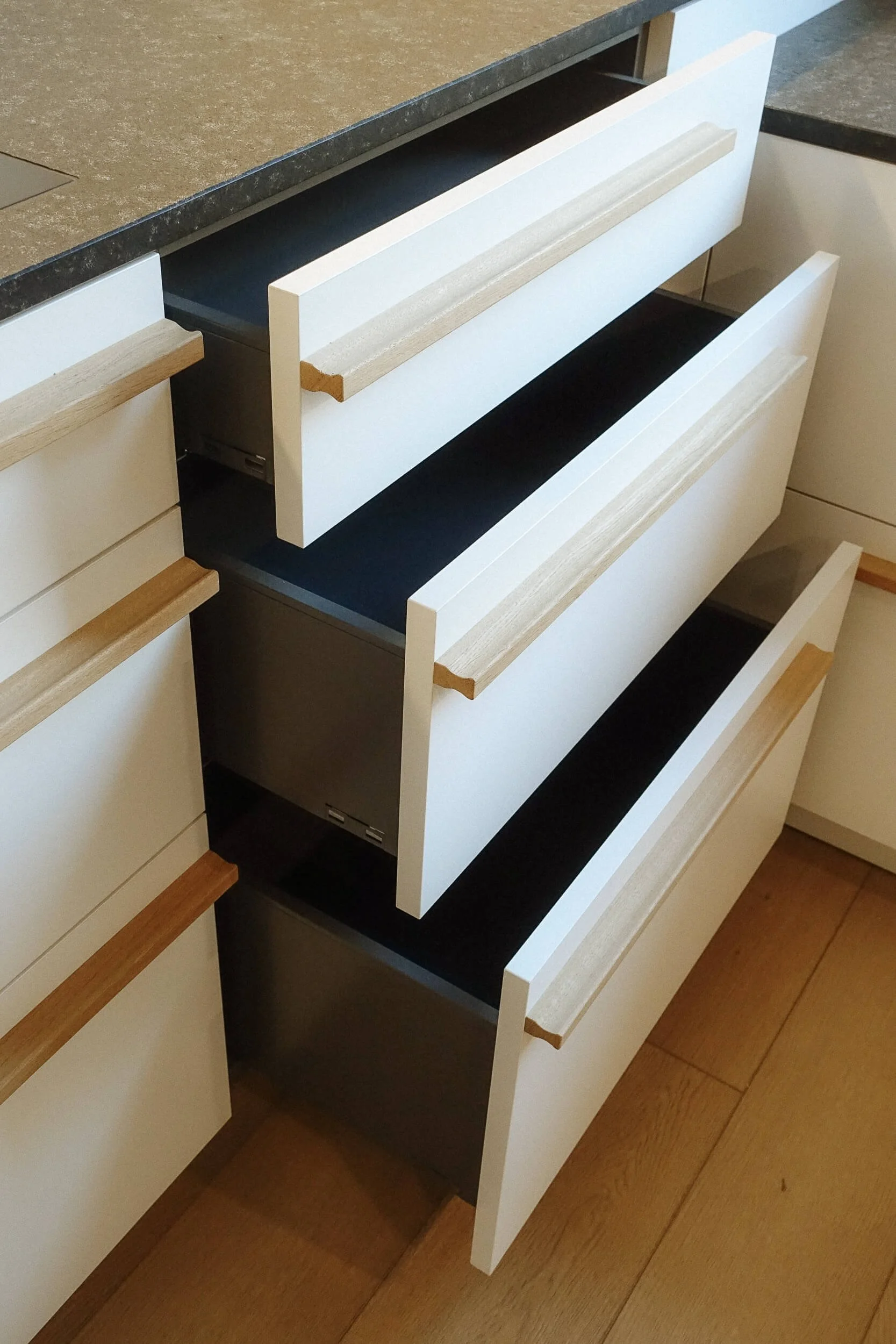 Offene Schubladen in einer weißen Küchenschublade mit Holzgriffen, oberhalb eine schwarze Arbeitsfläche.