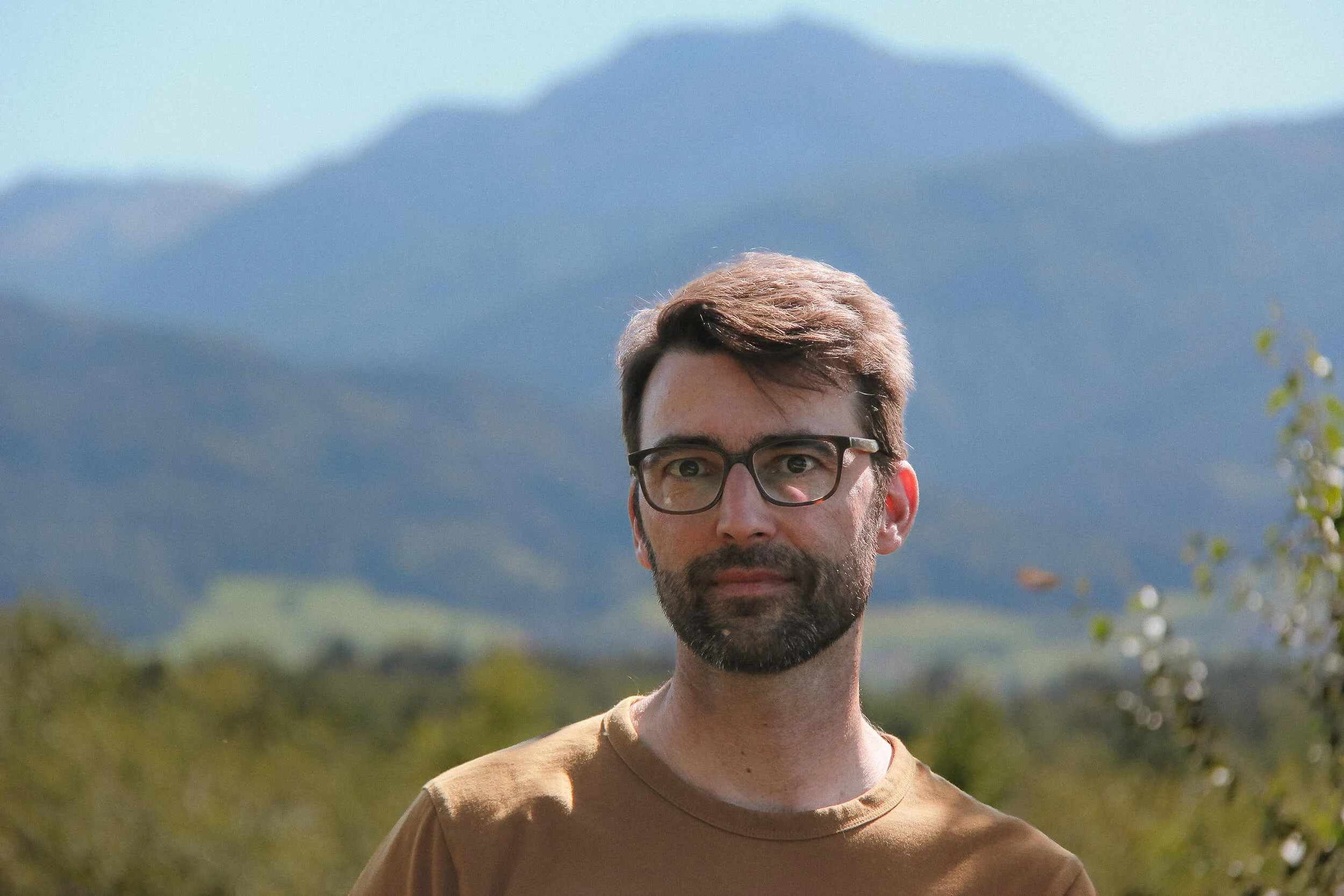 Porträt eines Mannes mit Brille im Freien vor Bergen, mit grünem Wasser und Bäumen im Hintergrund.