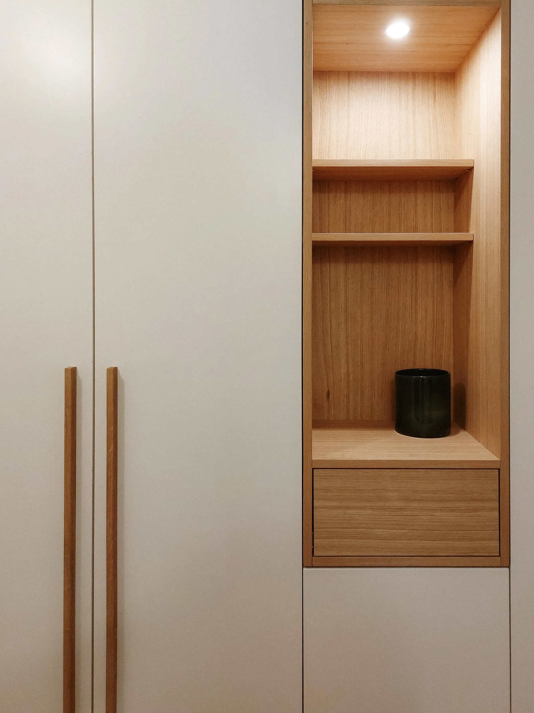 Ein moderner Einbauschrank mit weißen Türen und Holz-Details, darunter eine offene Nische mit zwei Ablagebrettern und einem schwarzen Behälter.