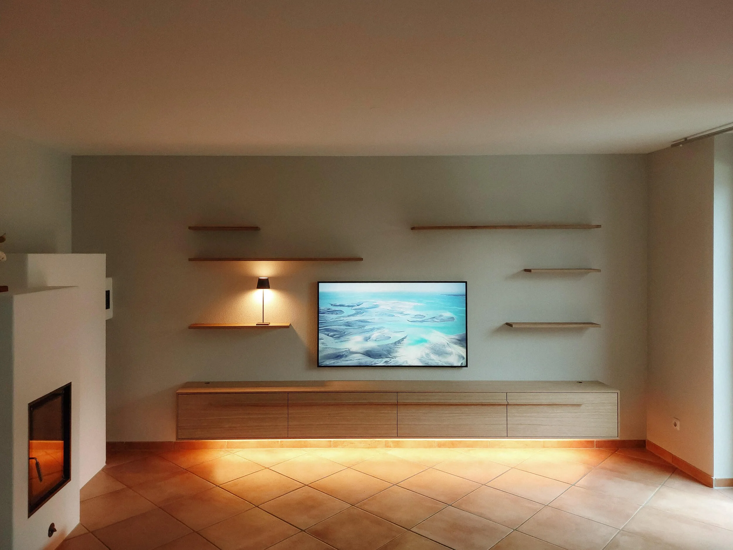 Innenaufnahme eines Wohnzimmers mit Wandregalen, einem Fernseher und warmem Bodenlicht.