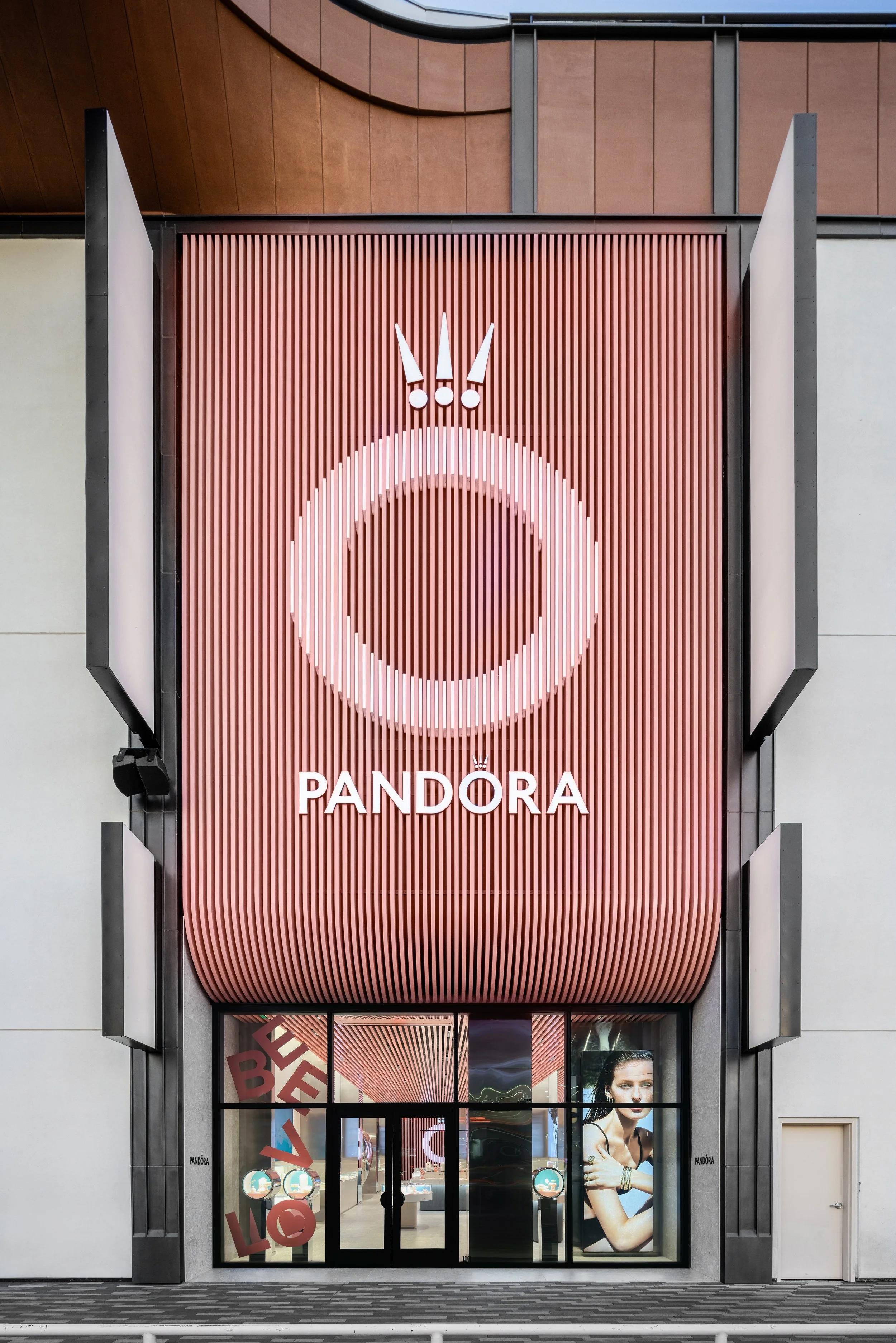 Exterior of a Pandora jewelry store with a large pink and white logo on a vertical brown ribbed backdrop, glass entrance doors, and promotional posters inside.