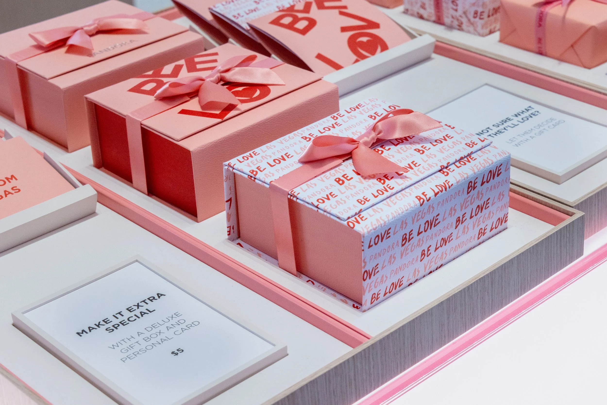 Multiple pink gift boxes with bows, some wrapped in 'Be Love' patterned paper, displayed on a store shelf, with a sign describing a special gift box and personal card for $5.