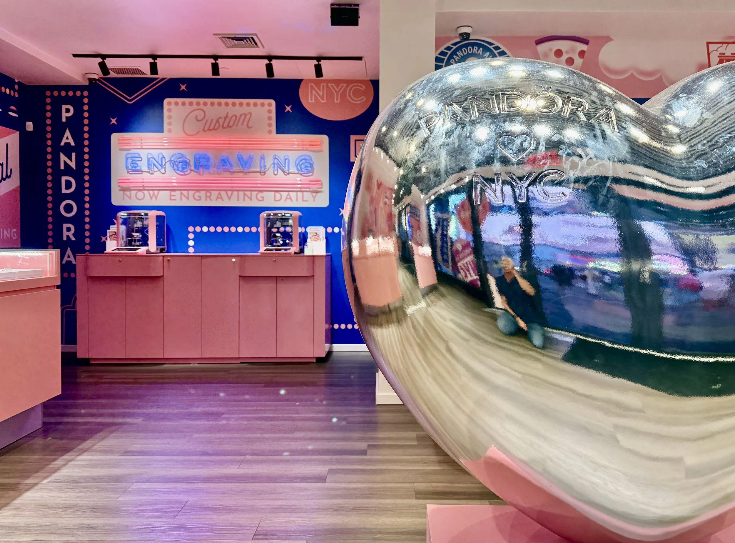 A display of engraved jewelry at Pandora store with a large reflective silver sphere in the foreground, and a pink counter in the background against a blue wall with neon signs reading 'Engraving' and 'Now Engraving Daily'.