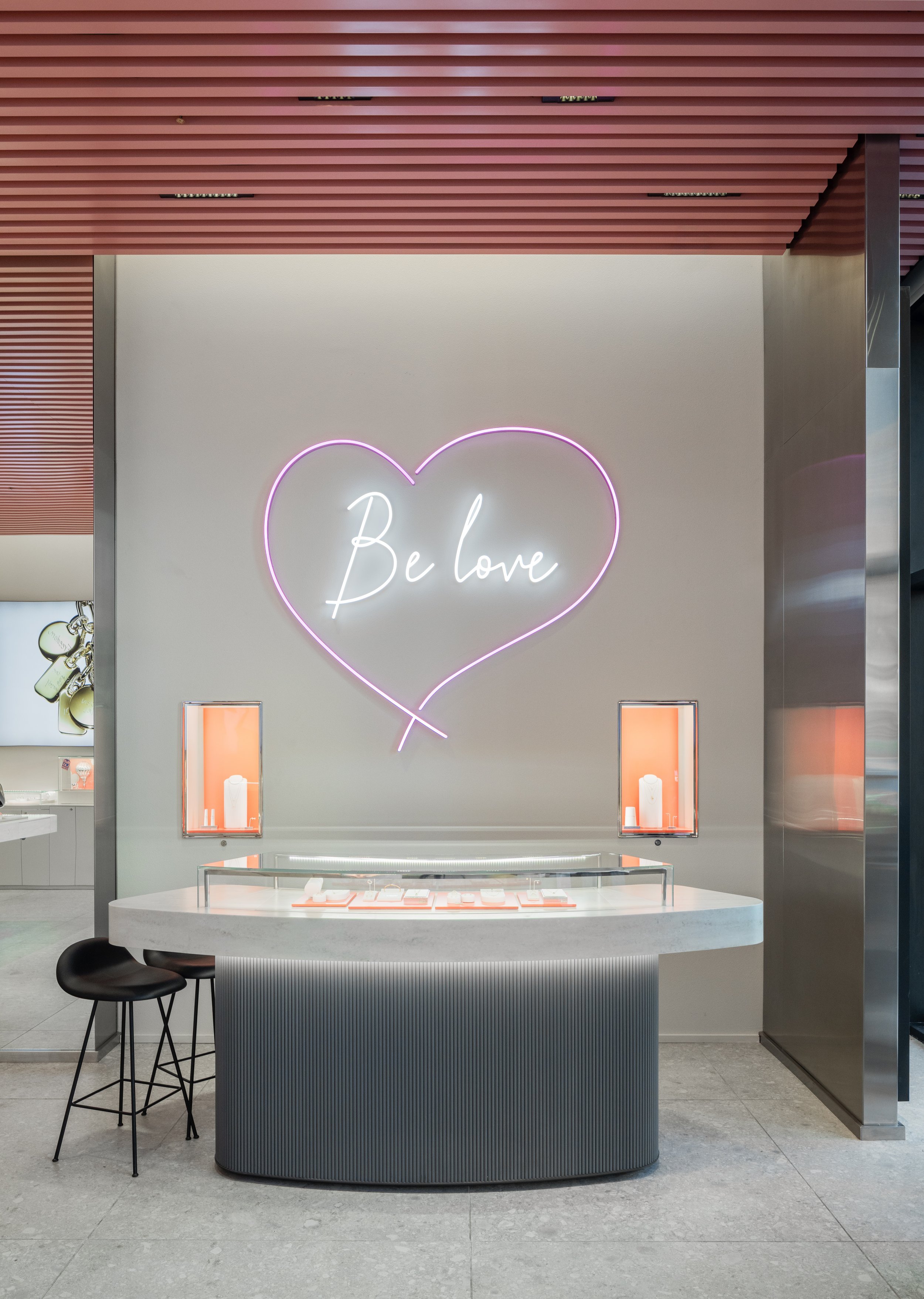 Neon sign with the words "Be love" inside a pink heart outline on a white wall, above a white display table with jewelry, in a modern retail store.