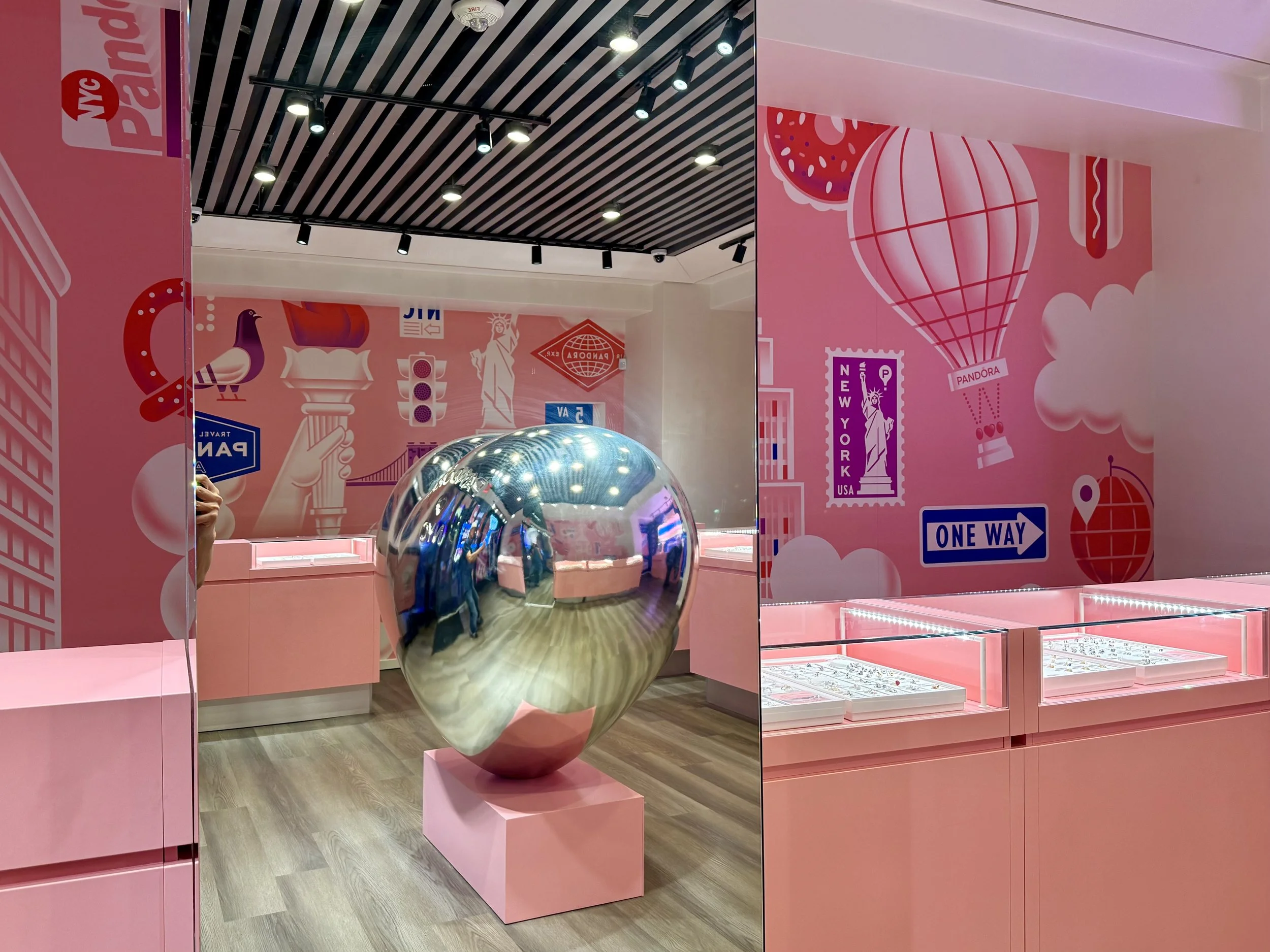 A pink-themed retail display featuring a large reflective metallic apple sculpture, glass display cases, and wall art with travel and city imagery including the Statue of Liberty, New York signs, and a hot air balloon.