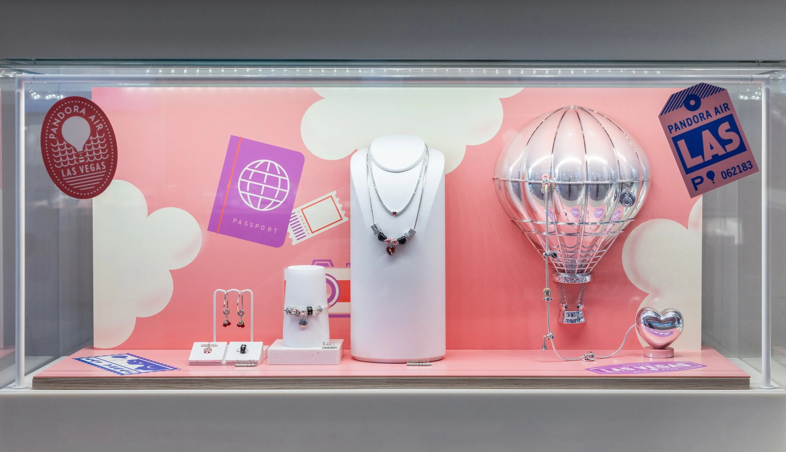 Jewelry display case with necklaces, earrings, and a bracelet on a pink background featuring travel-themed illustrations like a passport and an hot air balloon.