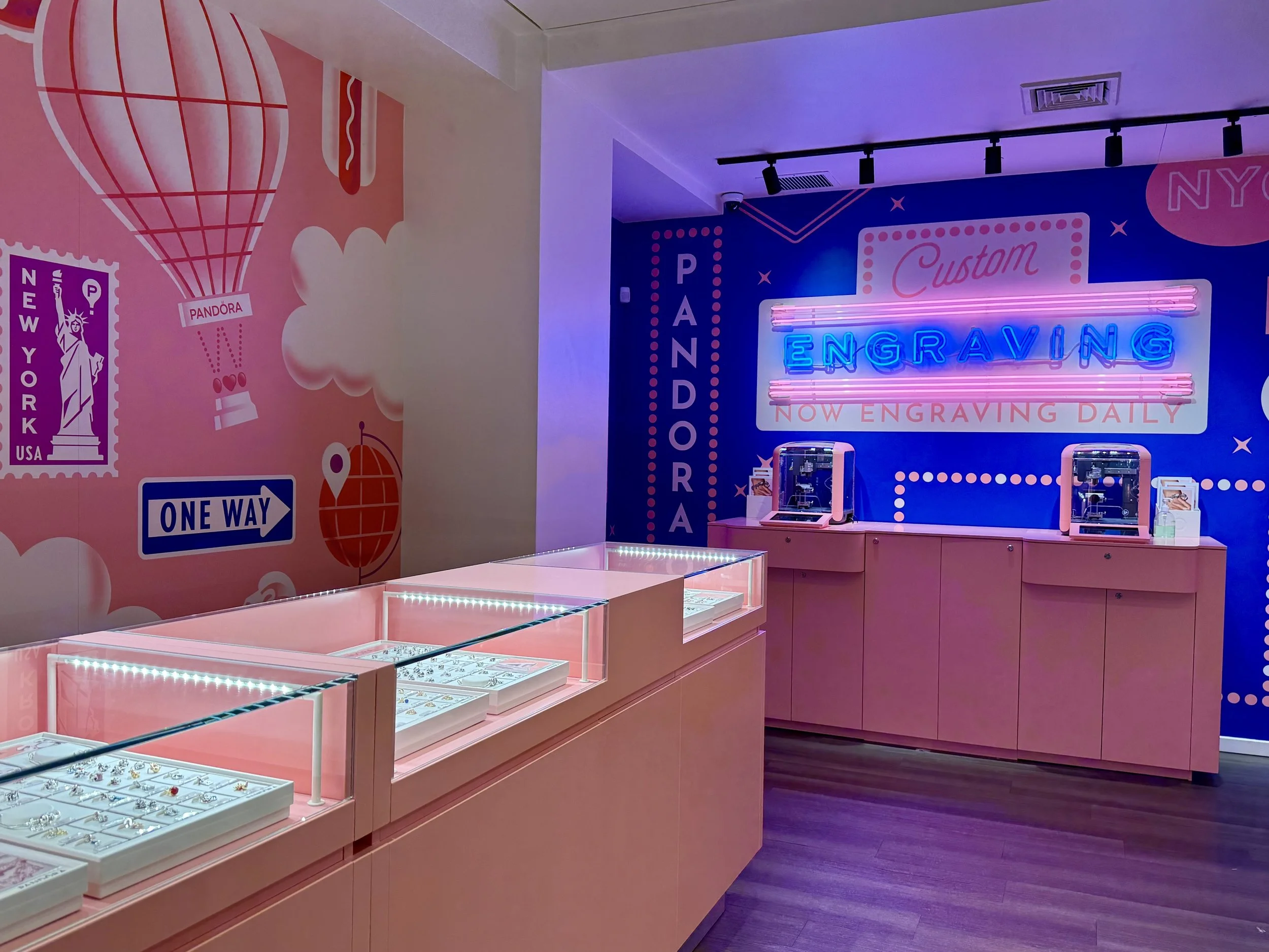 A jewelry store display with pink lit glass cases showcasing rings, with a pink and blue wall behind that features neon signs reading 'Engraving' and 'Now Engraving Daily'.