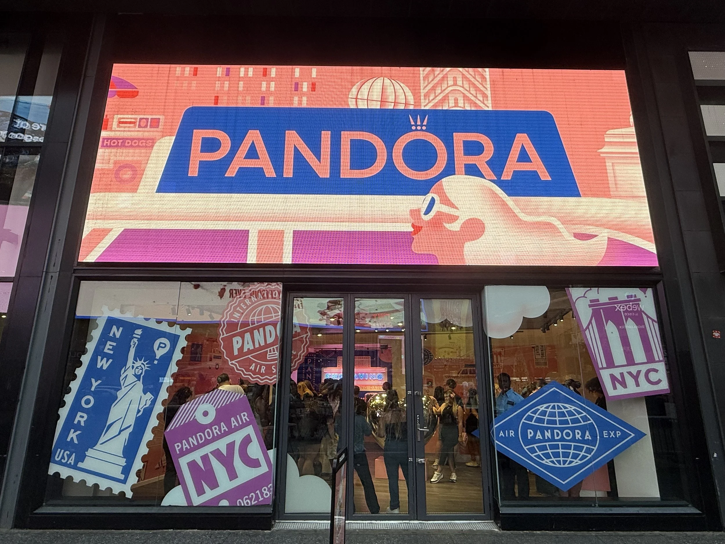 Storefront of Pandora with large digital sign and window display featuring various Pandora-branded graphics and NYC-themed stamps, with people inside visible through the glass.