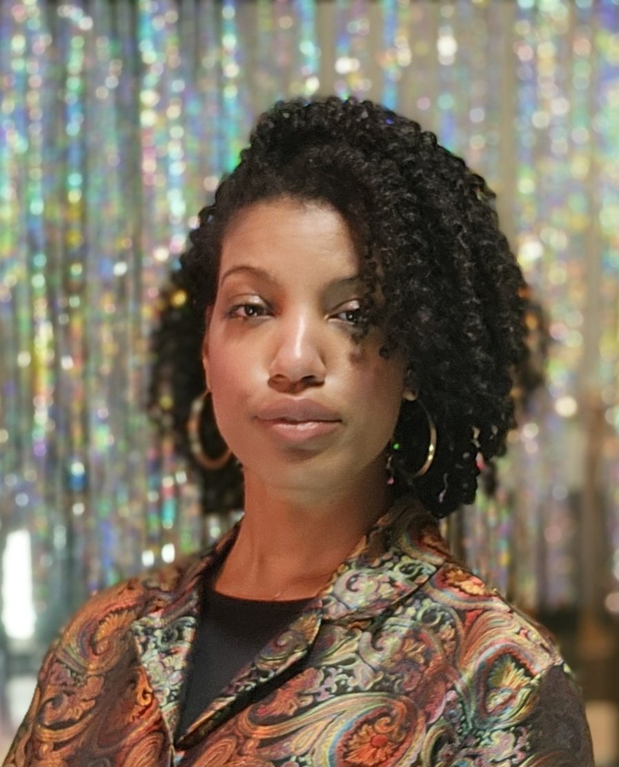 Portrait of the artist, a beautiful young woman with curly black hair, hoop earrings, wearing a satin multicolored paisley-patterned jacket, standing in front of a shiny, iridescent backdrop.