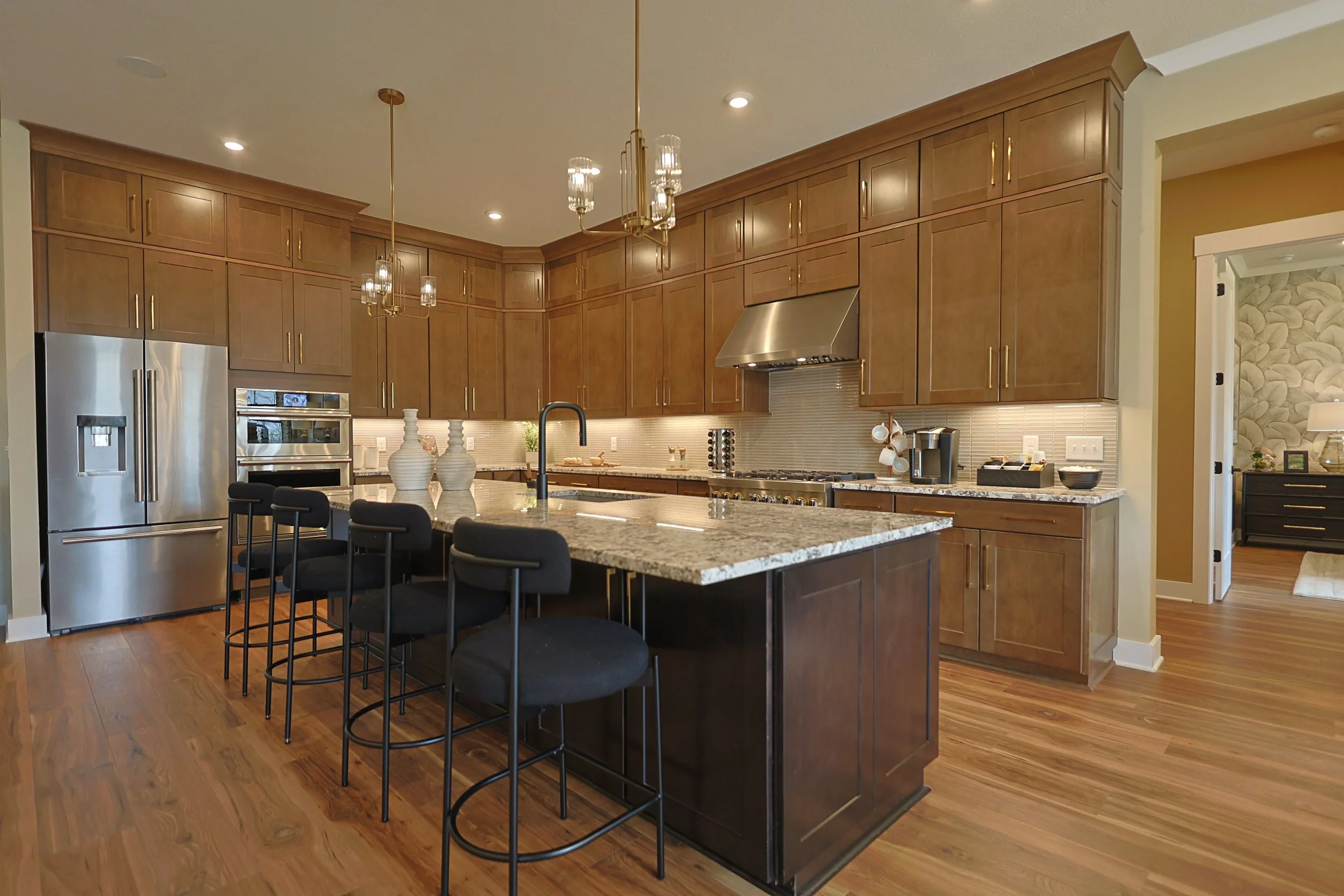Modern kitchen with wooden cabinetry, stainless steel appliances, a large granite island, black chairs, pendant and chandelier lighting, and hardwood flooring.