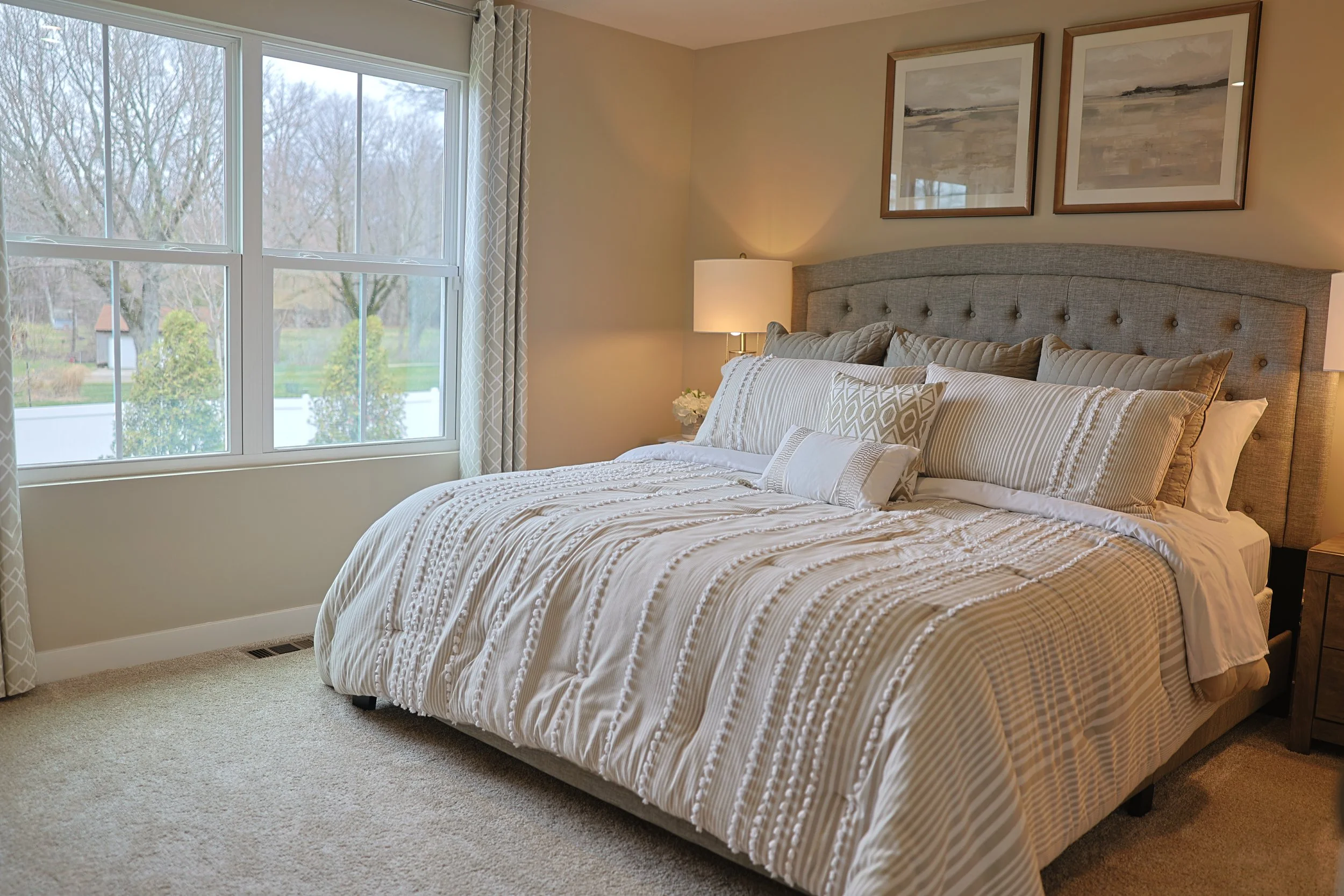 A neatly made bed with a gray upholstered headboard, multiple decorative pillows, and a cream-colored striped bedspread in a cozy bedroom with a large window showing an outdoor scene with trees and a small shed.