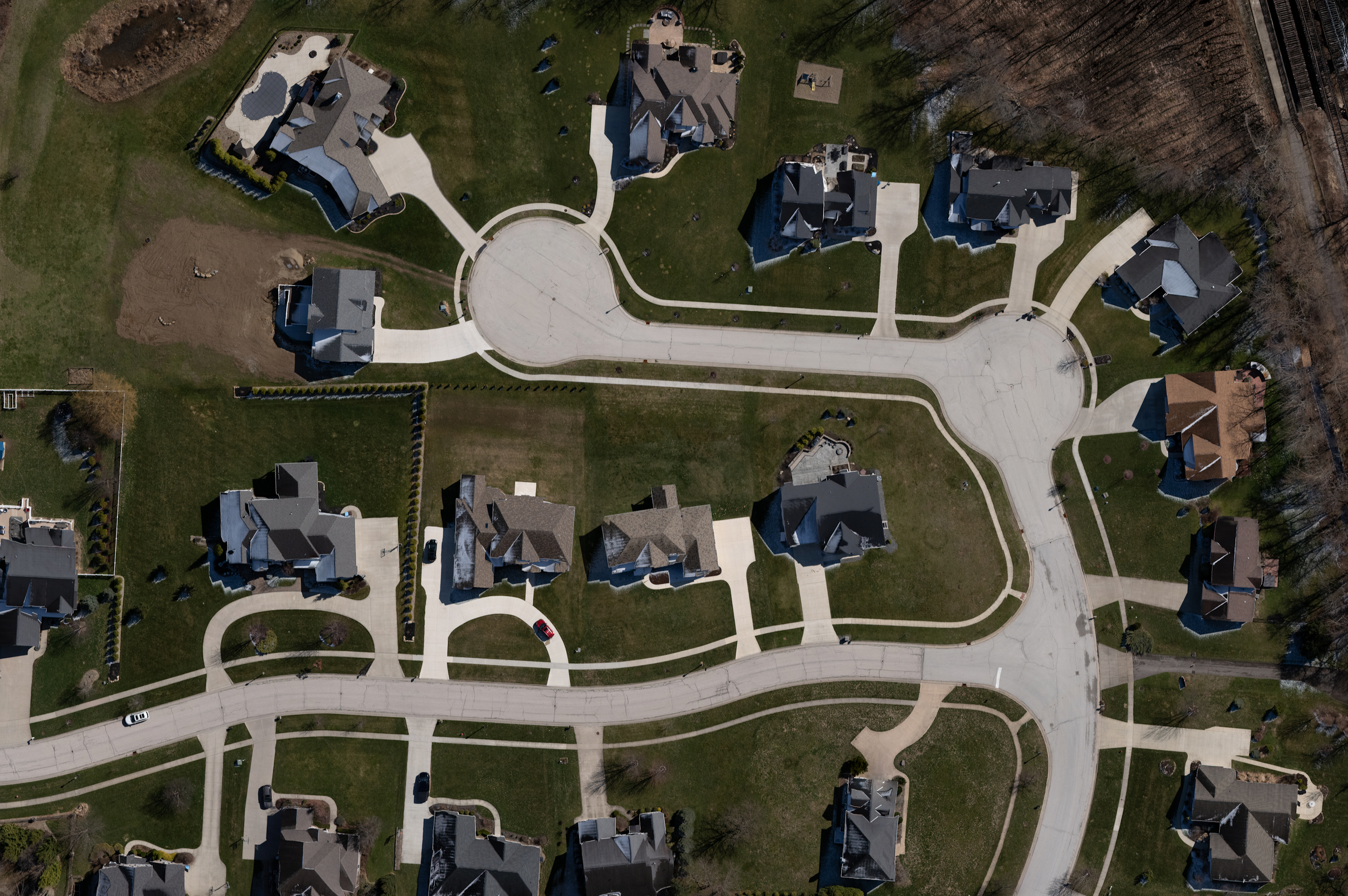 Aerial view of a residential neighborhood showing houses, green lawns, curved streets, and a central cul-de-sac, with some trees and a wooded area on the top right.