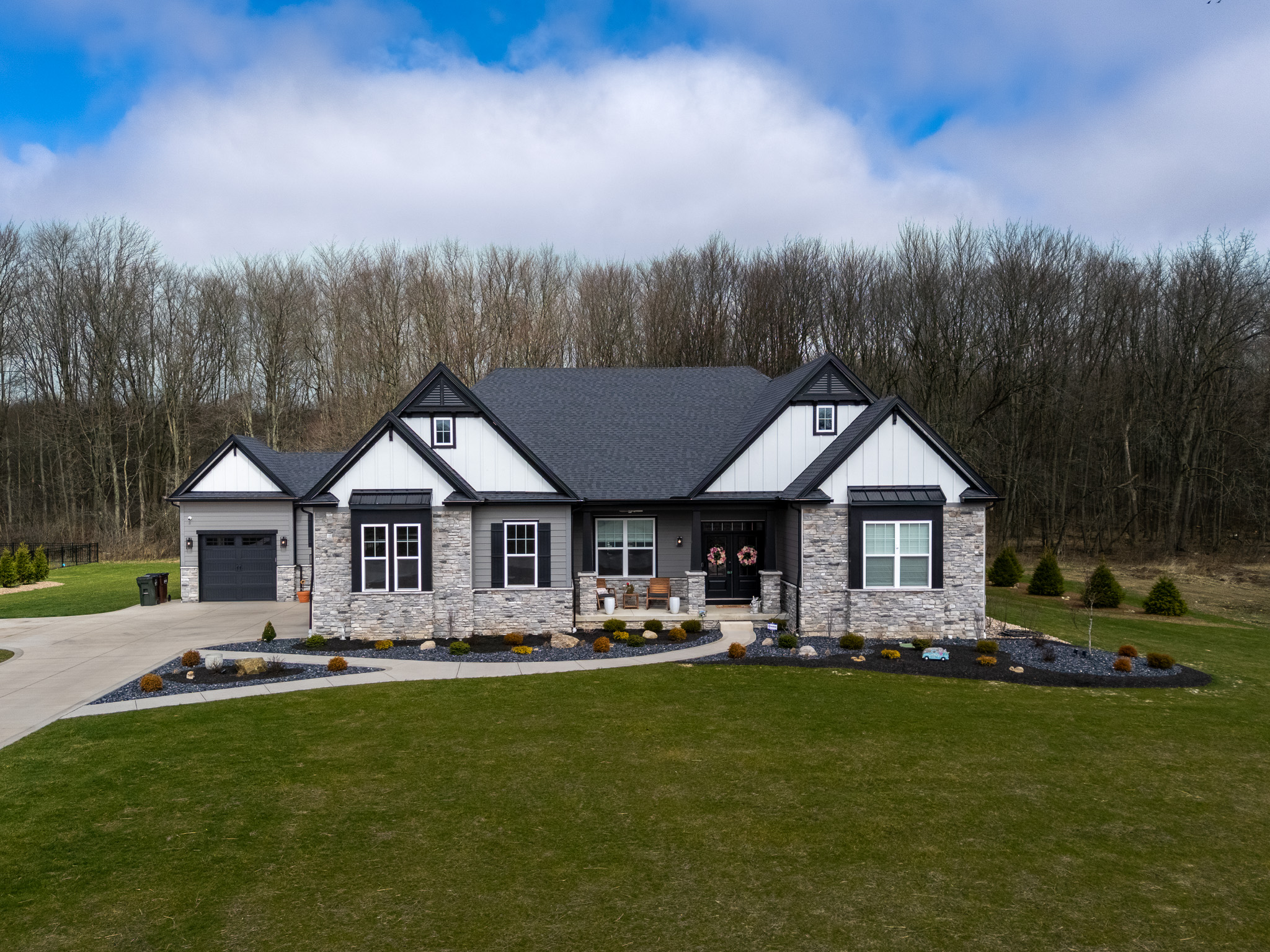 Large modern house with gray stone and white siding, black accents, and a black garage door, surrounded by a well-manicured lawn and landscaped garden beds with small bushes.
