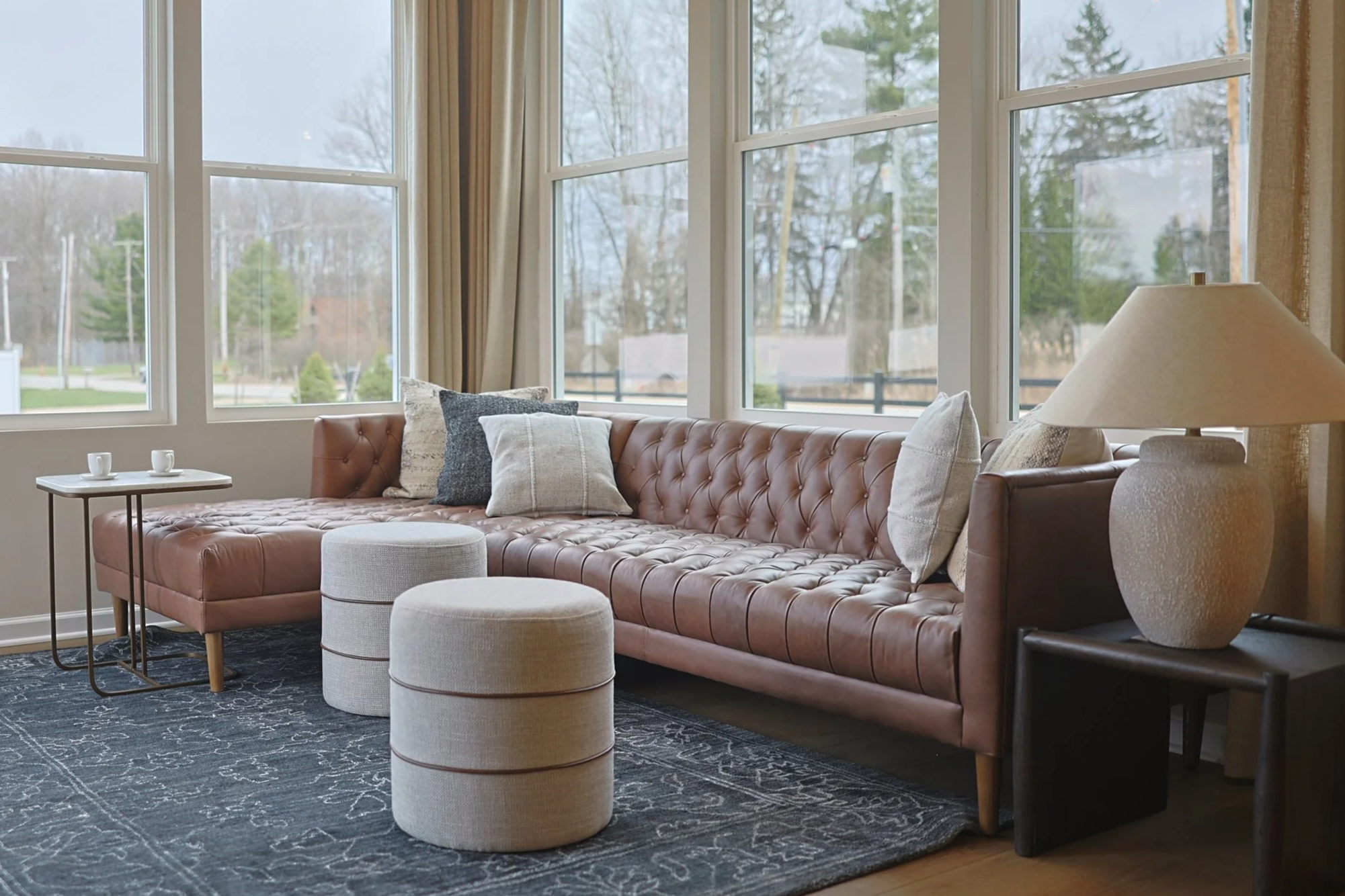 Living room with a caramel leather tufted sectional sofa, beige and gray pillows, two round beige ottomans, a dark side table with a large beige lamp, a patterned blue area rug, and large windows with beige curtains overlooking a suburban neighborhoo
