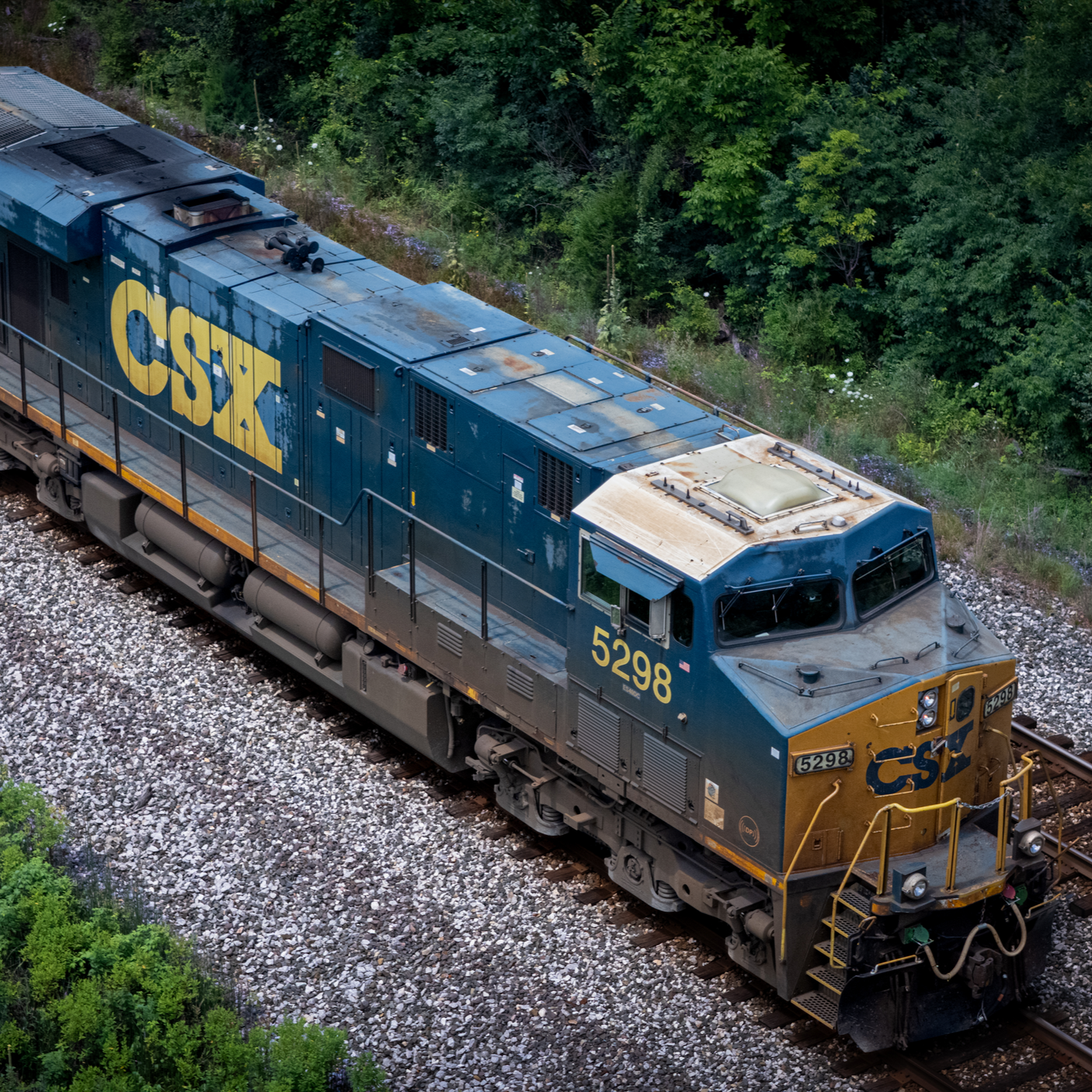 An aerial view of a CSX freight train engine traveling on railway tracks through a green, wooded area.