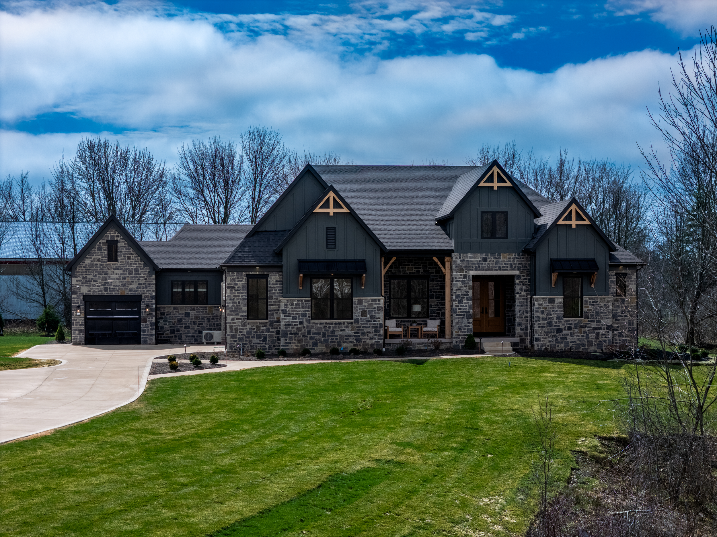 A modern, two-story house with stone and dark siding exterior, front porch, and attached garage, surrounded by a well-maintained lawn with trees and a driveway.