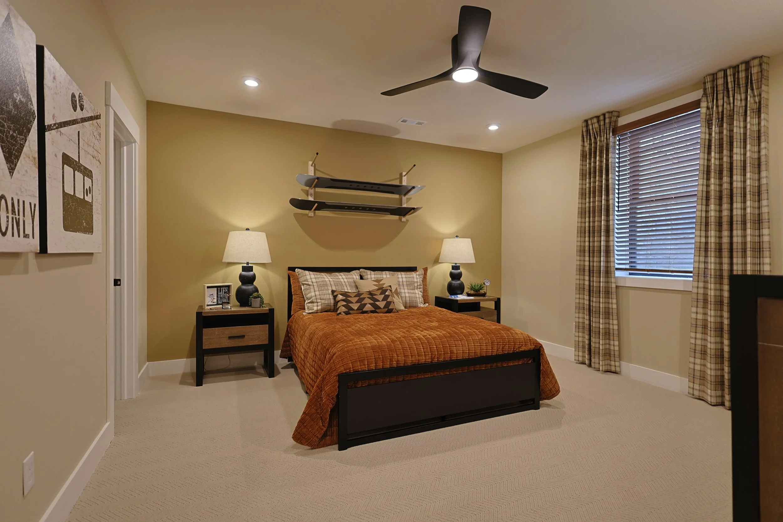 A bedroom with beige walls, a black bed frame, and an orange bedspread. There are two black nightstands with lamps, and a window with plaid curtains. A skateboard shelf is mounted above the bed.