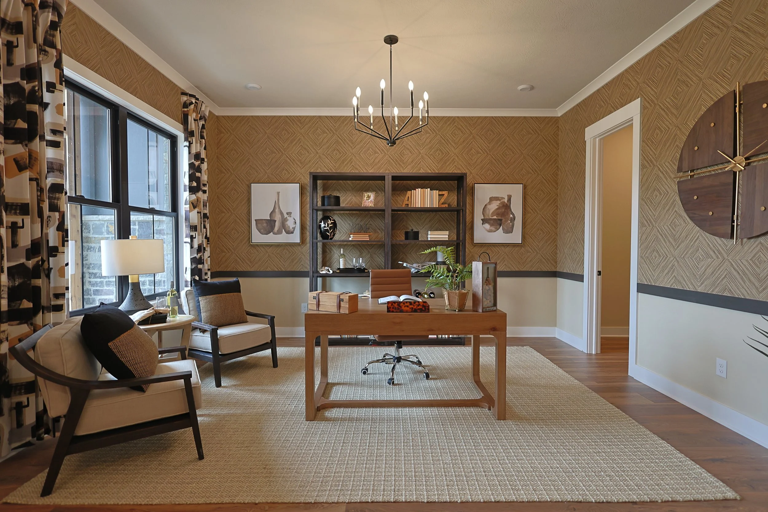 A well-lit modern office with a wooden desk and a gray office chair, flanked by two beige armchairs with dark wooden frames, positioned on a light-colored rug. A black shelving unit with books and decorative items is against the patterned brown wallp
