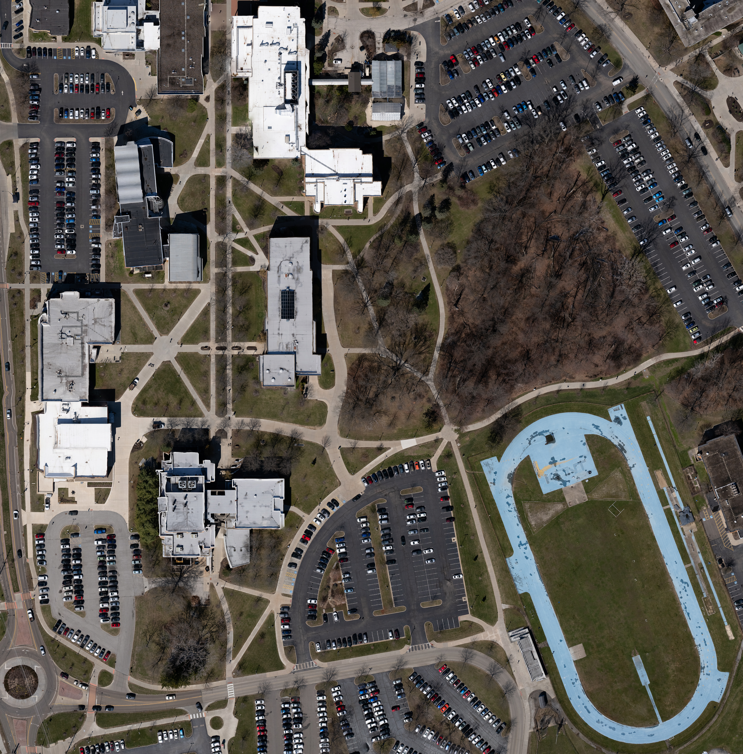 An aerial view of a university campus with multiple white buildings, parking lots, a park with trees, and a blue running track.