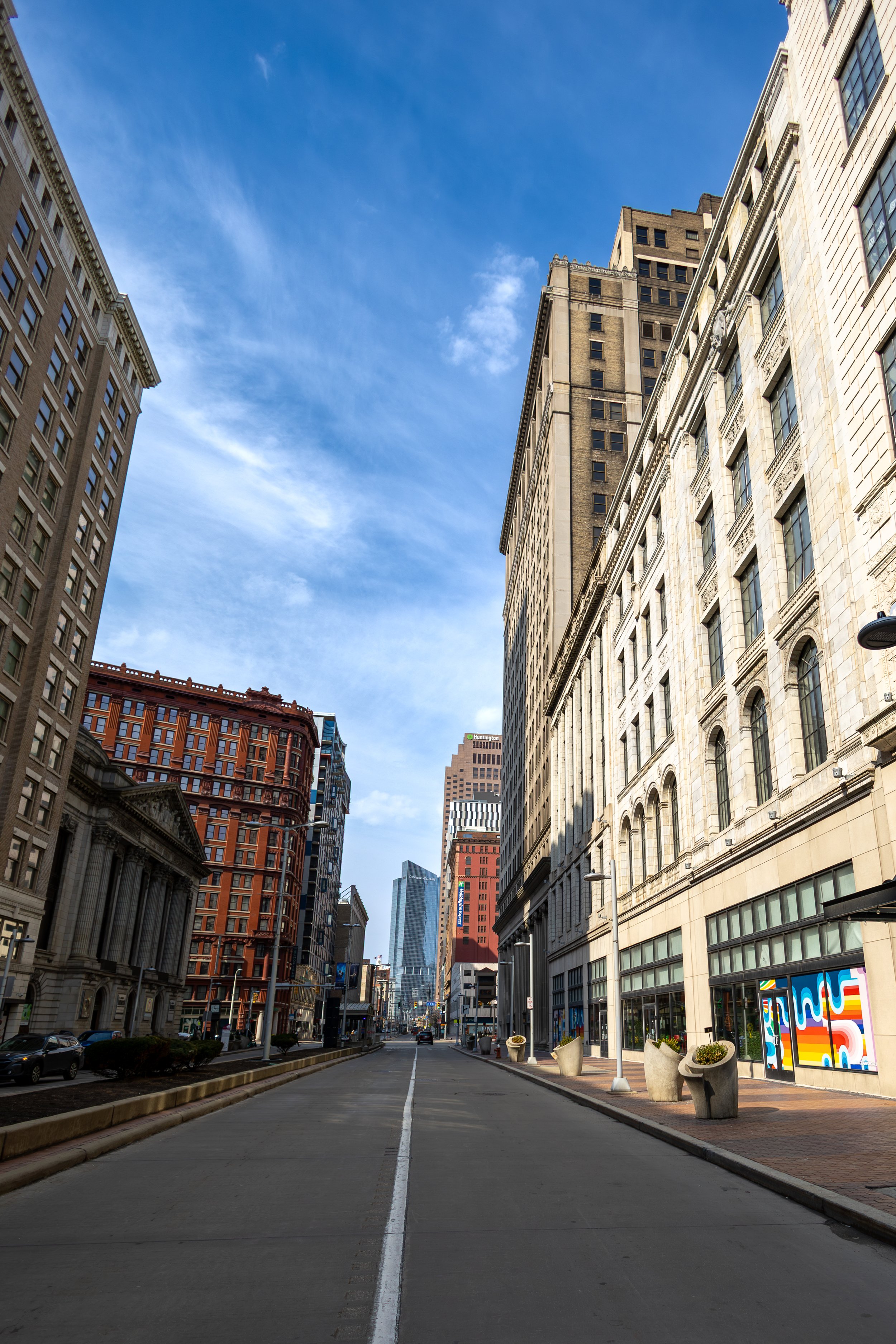 Empty city street lined with tall historic buildings under a partly cloudy blue sky.