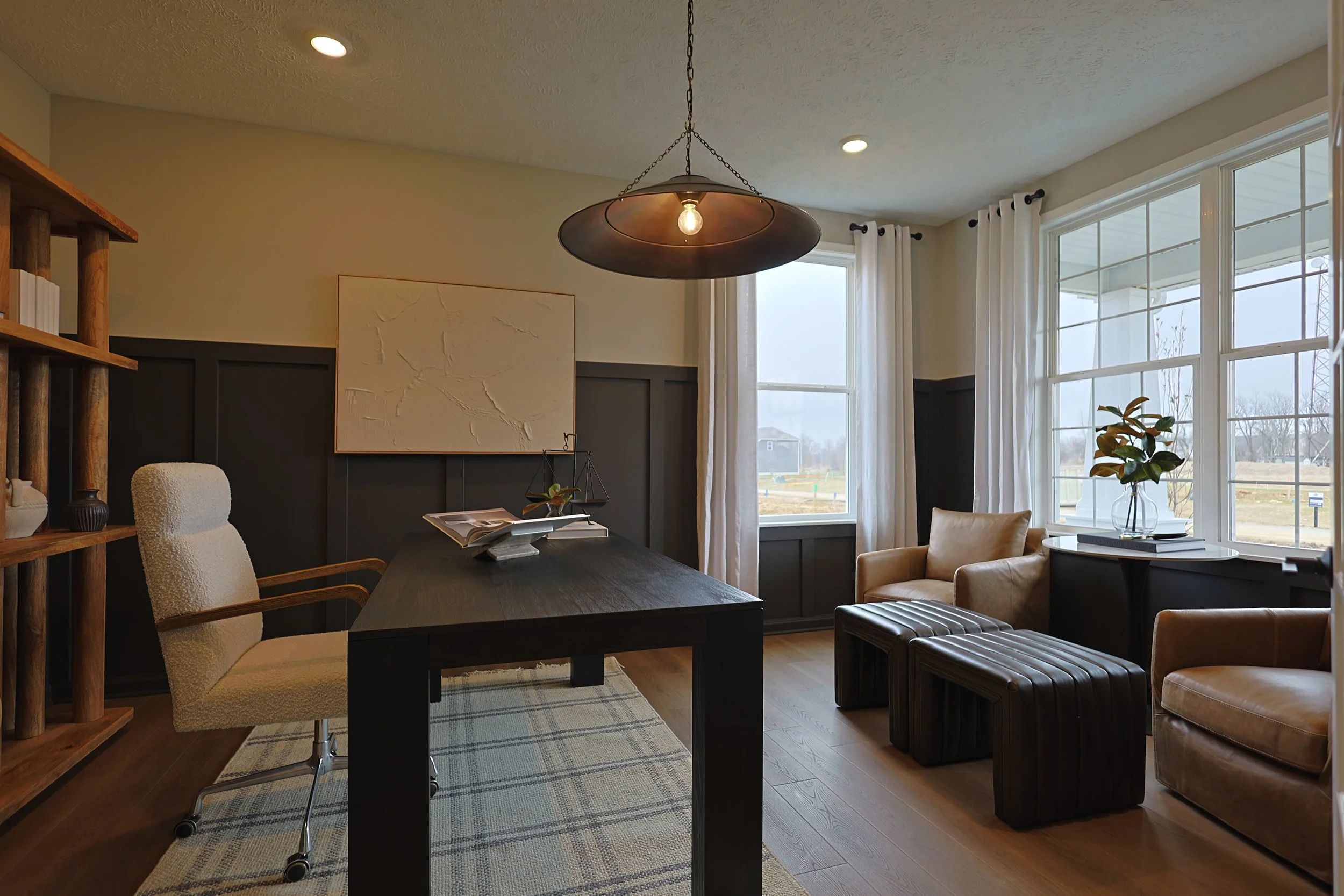 A cozy home office with a black wooden desk, a beige cushioned office chair, two beige armchairs, a small black ottoman, large windows with white curtains, a wooden bookshelf, a modern hanging light fixture, and a potted plant on a table near the win