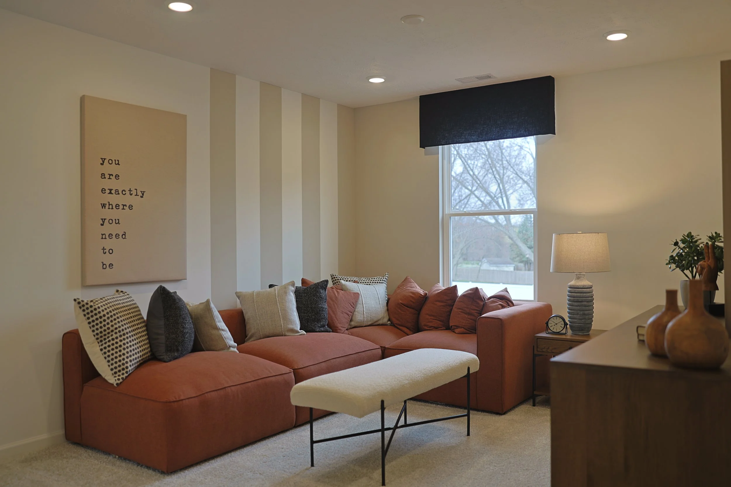 Living room with a burnt orange sectional sofa, a cream-colored bench, a table lamp, and pillows. There are vases and a small plant on a wooden sideboard, with a window showing trees outside. Wall art reads, 'you are exactly where you need to be.'