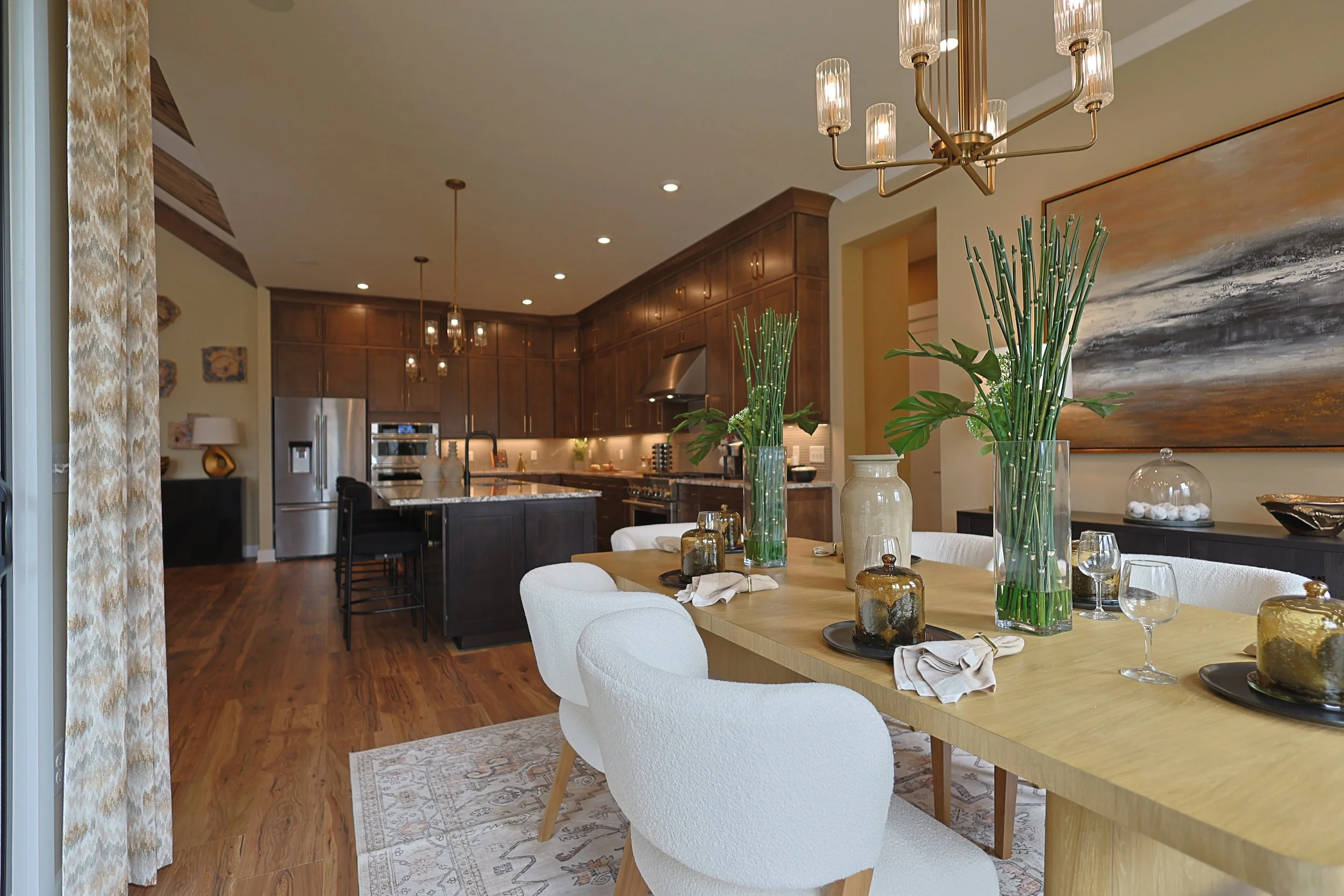 Open-concept dining and kitchen area with wooden flooring, white chairs around a wooden table decorated with large glass vases with green plants, set with cloth napkins, glasses, and jar containers. The kitchen features dark cabinets, a stainless ste