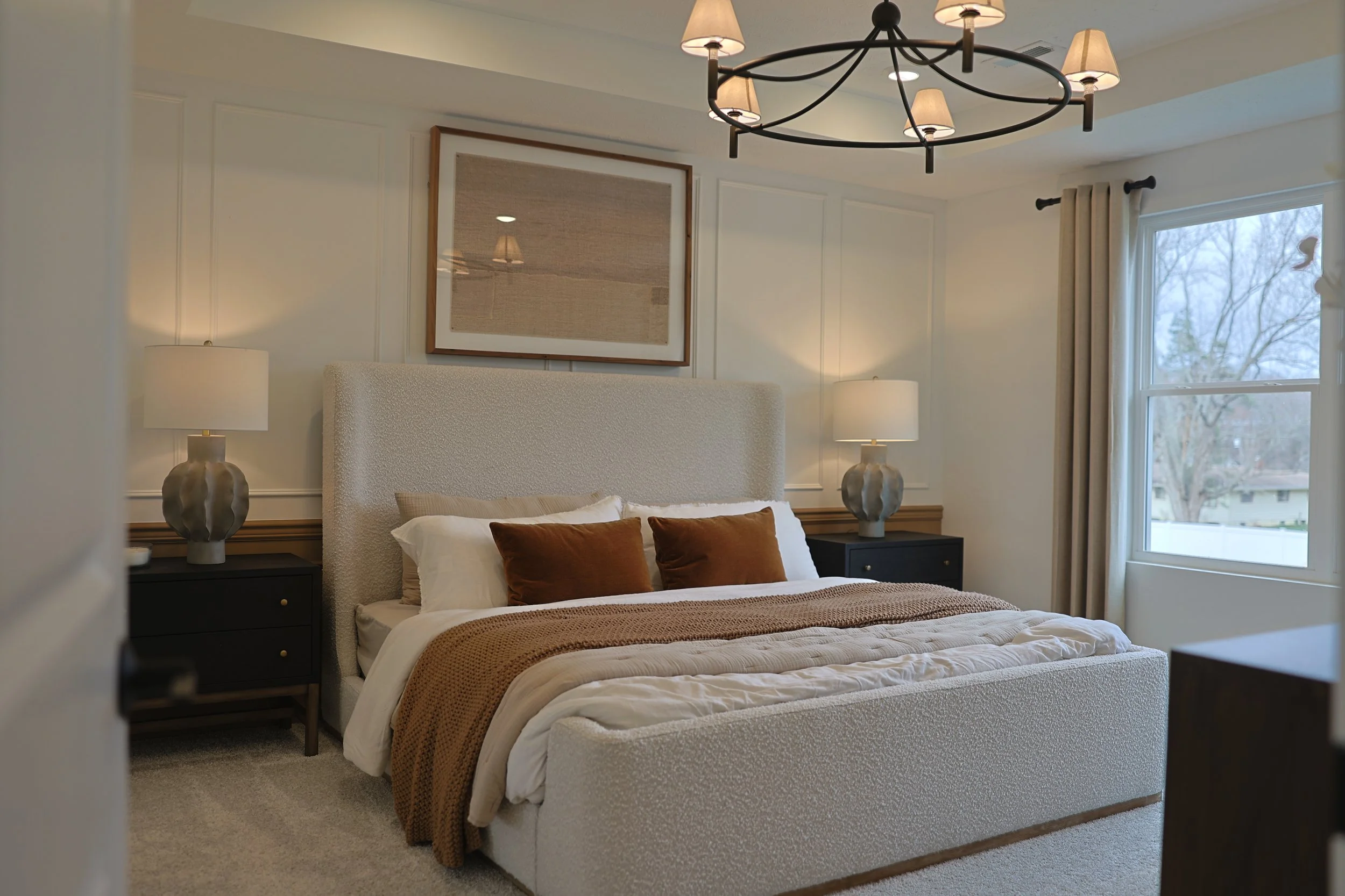 A neatly made bedroom with a king-size bed, beige upholstered headboard, white and brown bedding, and four decorative pillows, flanked by black nightstands with large gray lamps. There is a large window with beige curtains and a black chandelier hang