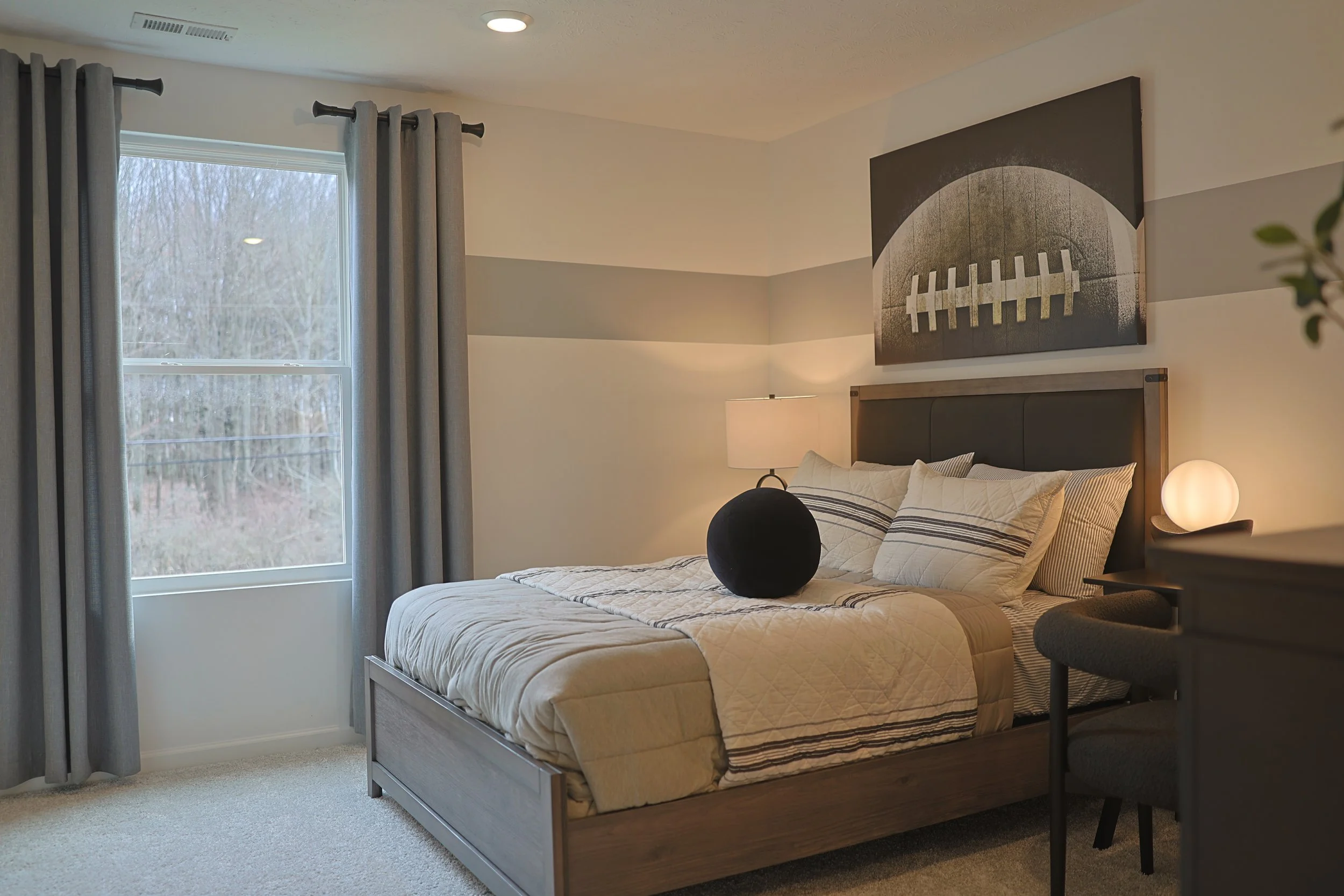 A neatly made bed with striped pillows in a modern bedroom with a large window, gray curtains, a painting of a football on the wall, and a minimalist nightstand with a spherical lamp.