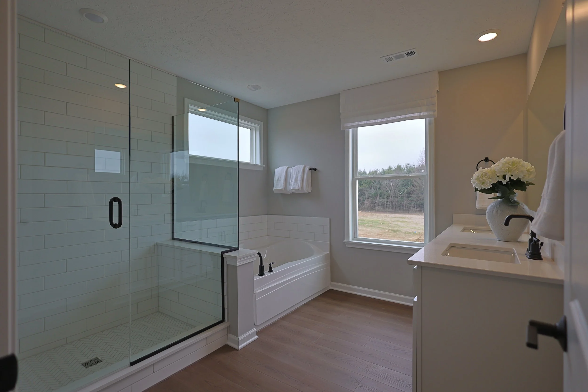 Bright bathroom with glass shower enclosure, white bathtub, double vanity with white countertop and sink, large window with outdoors view, small white towel on wall, vase with white flowers, hardwood floor, neutral wall colors.