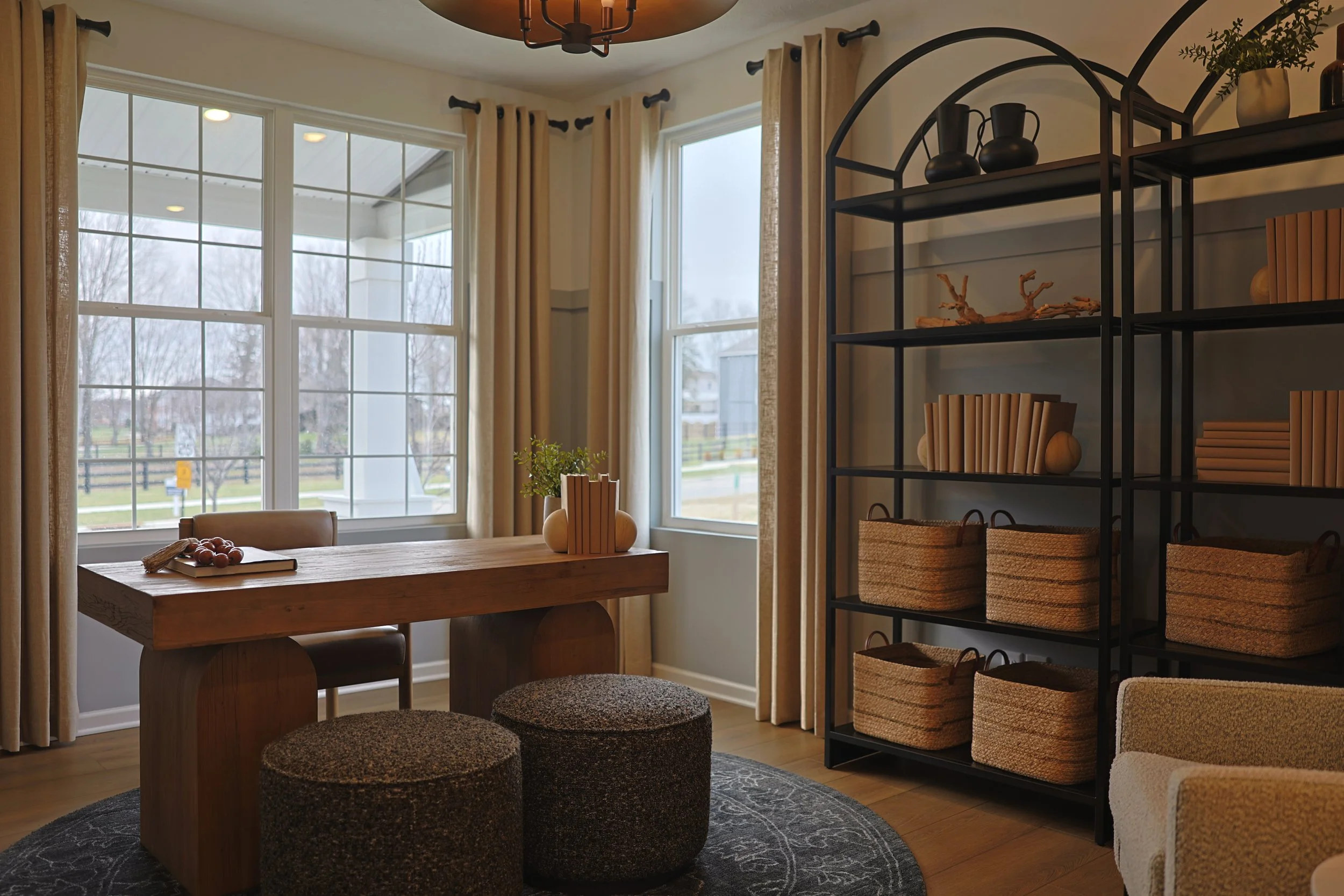 A cozy dining nook with a wooden table, two round ottomans, and a large window with beige curtains. There is a black metal shelf filled with baskets and decorative items, and a view of a backyard with trees outside.