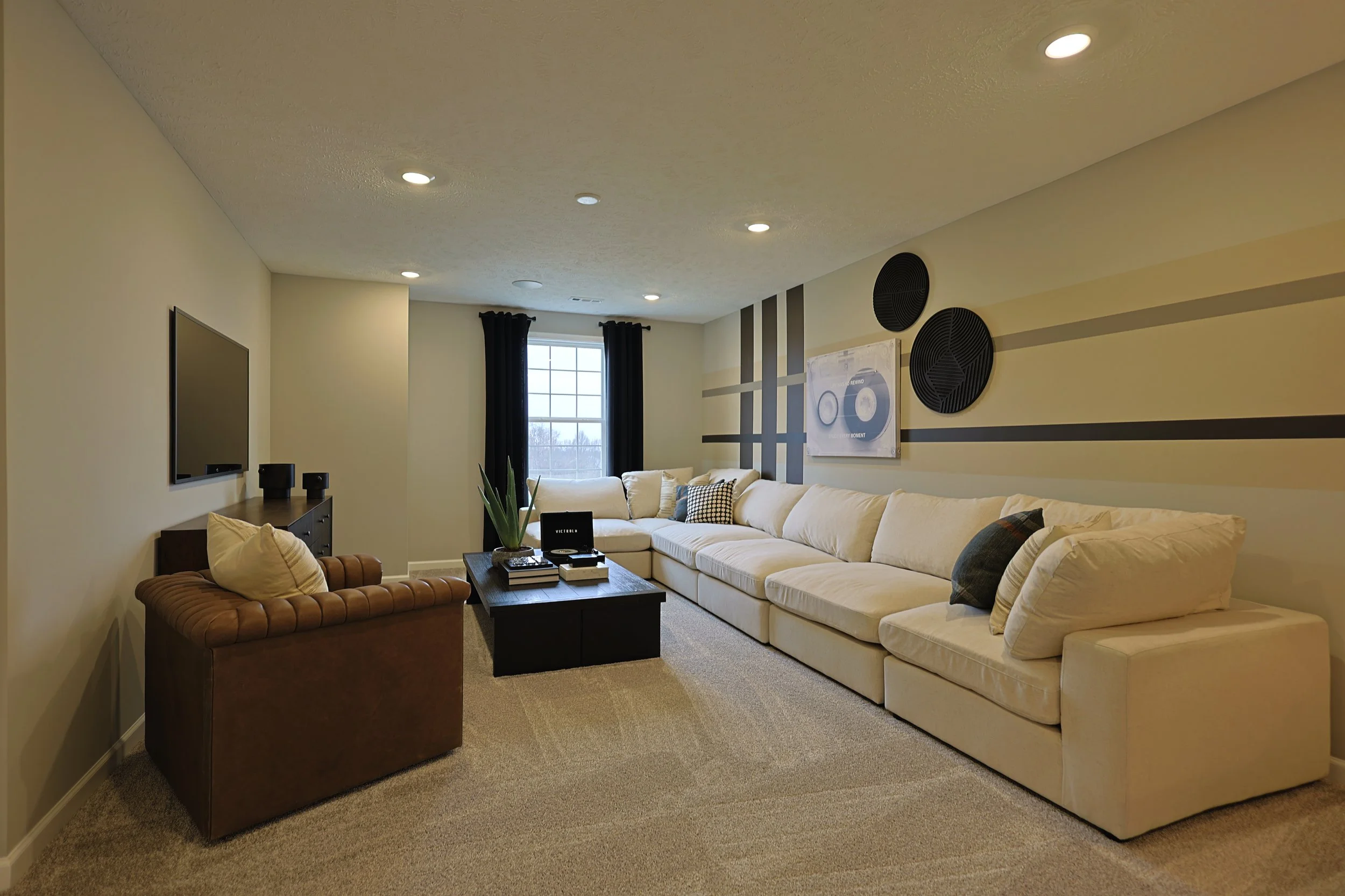 Living room with a beige sectional sofa, a brown armchair, a black coffee table, a wall-mounted TV, and a window with black curtains.